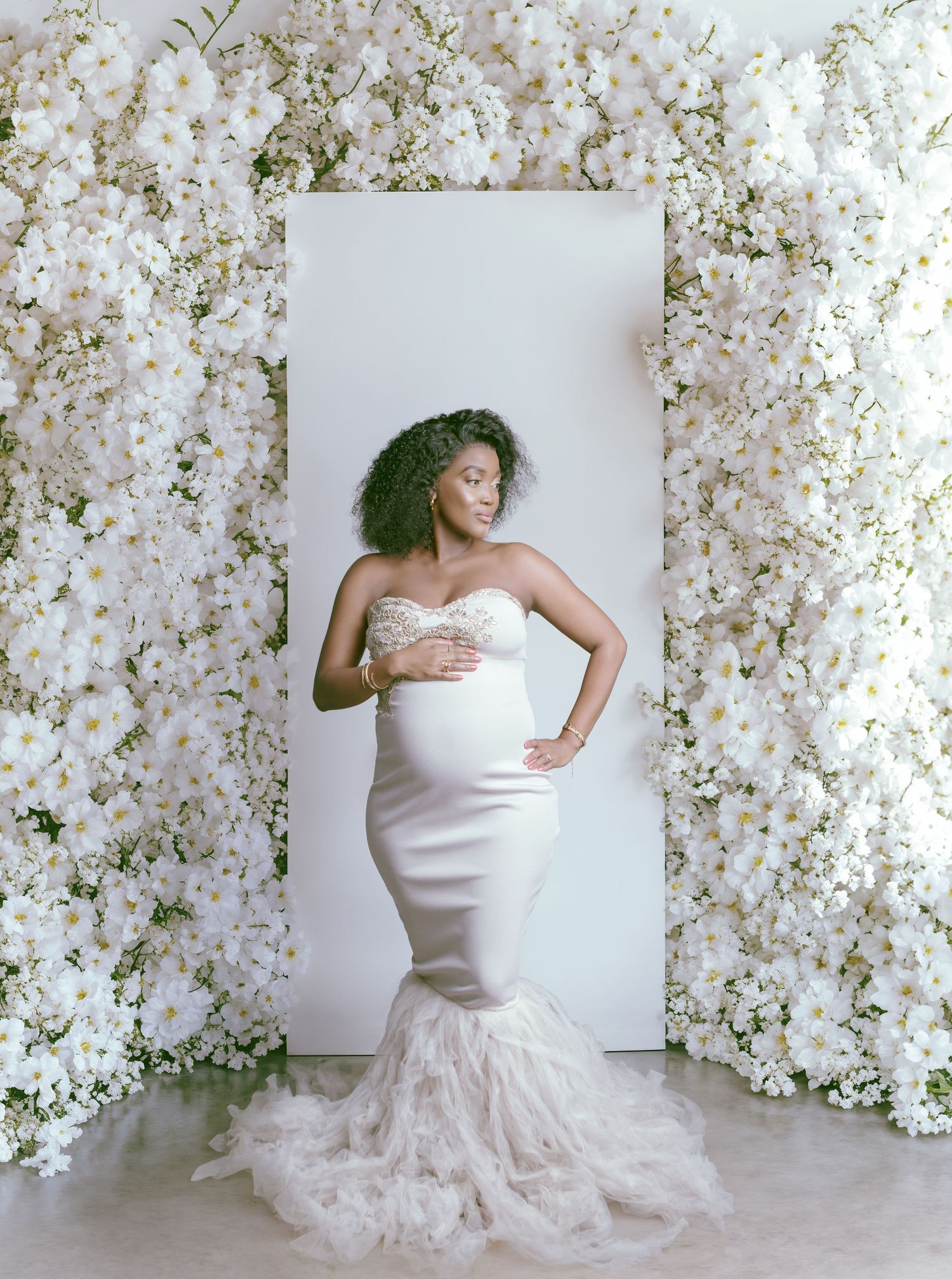 Elegant maternity portrait featuring a white mermaid-style gown with feathered hem surrounded by delicate white blossoms.