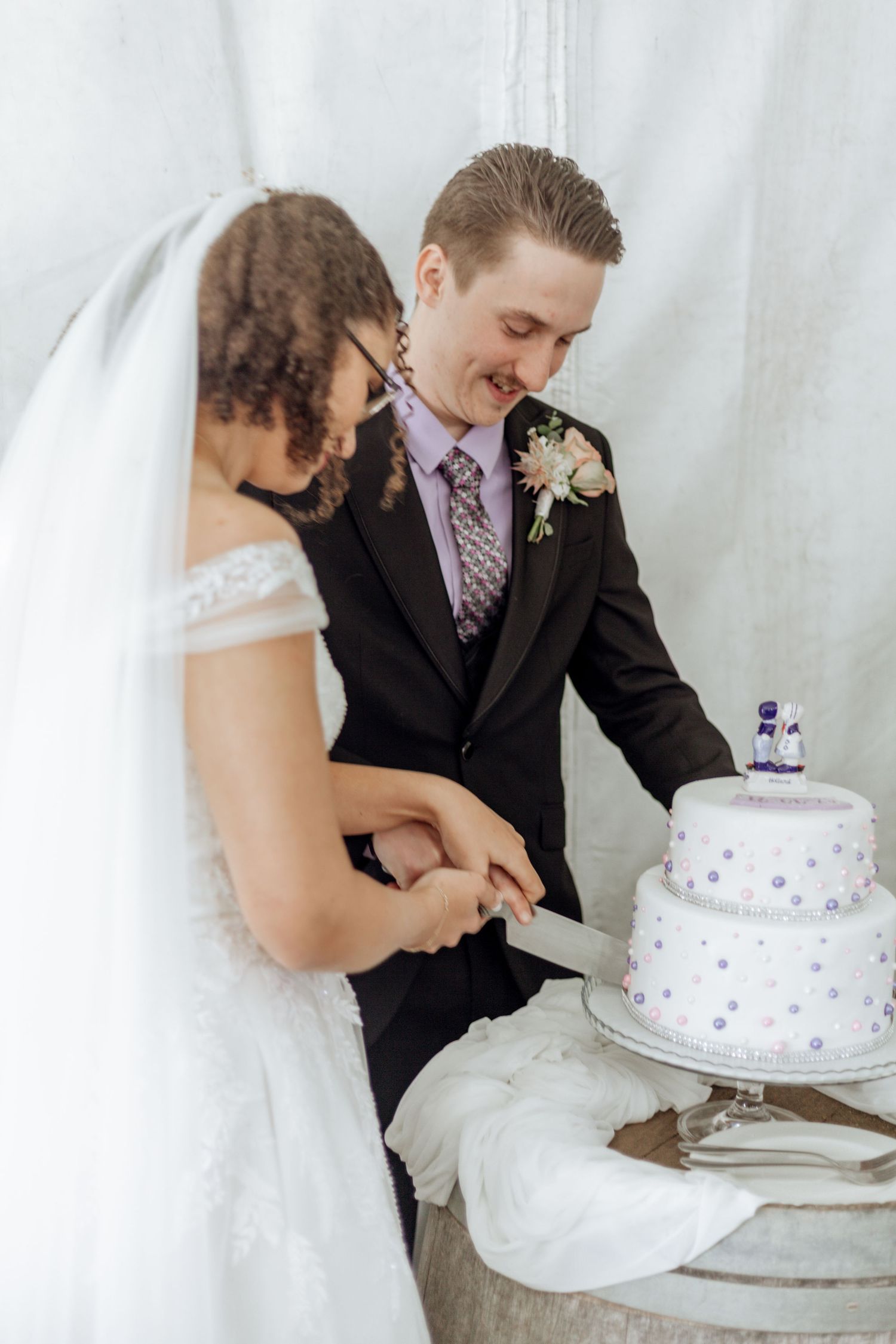 A couple cuts into their white and purple wedding cake together during their reception celebration.