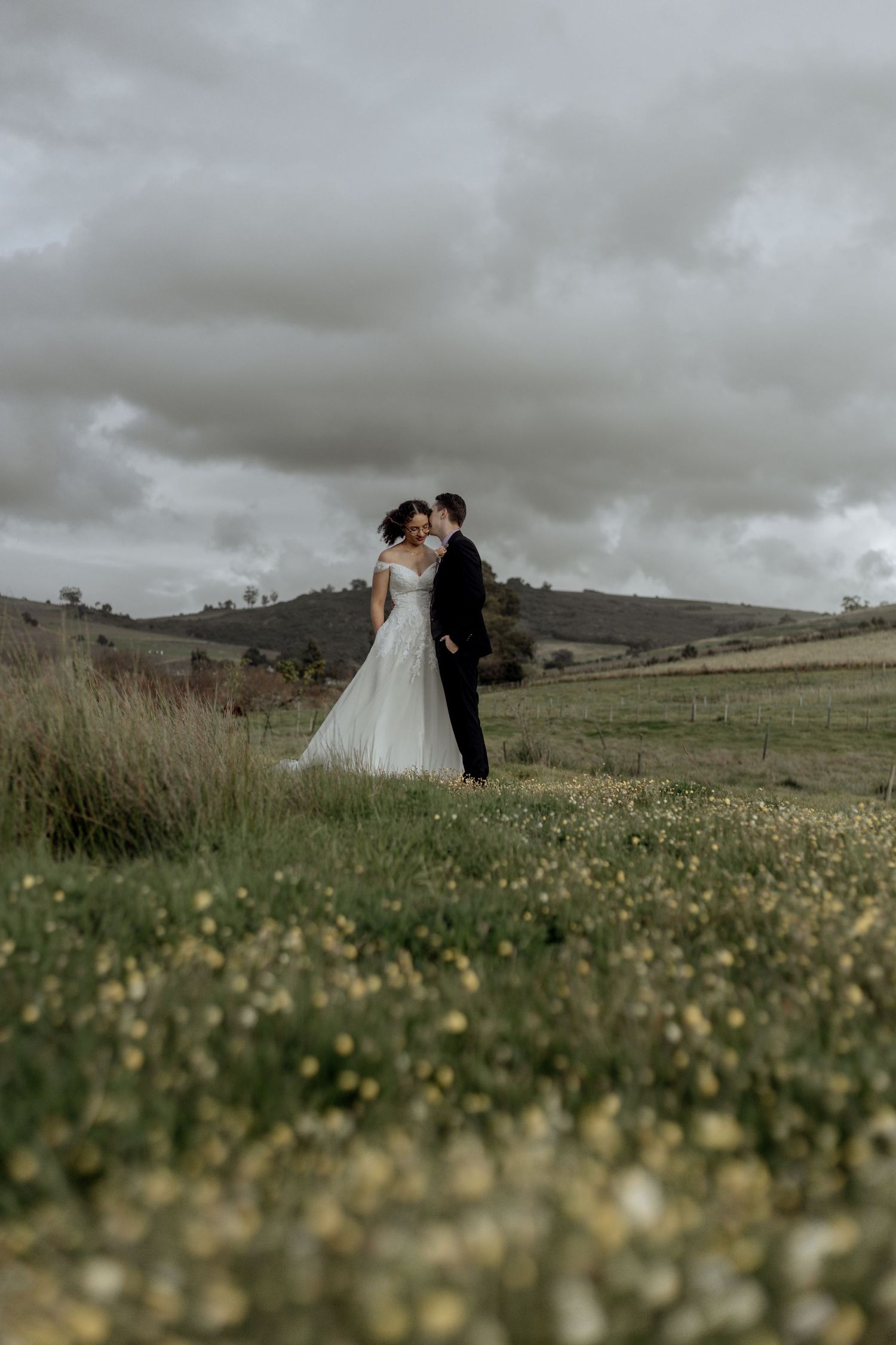Wide flowering meadow in overcast weather with distant couple embracing while silhouetted against moody sky.
