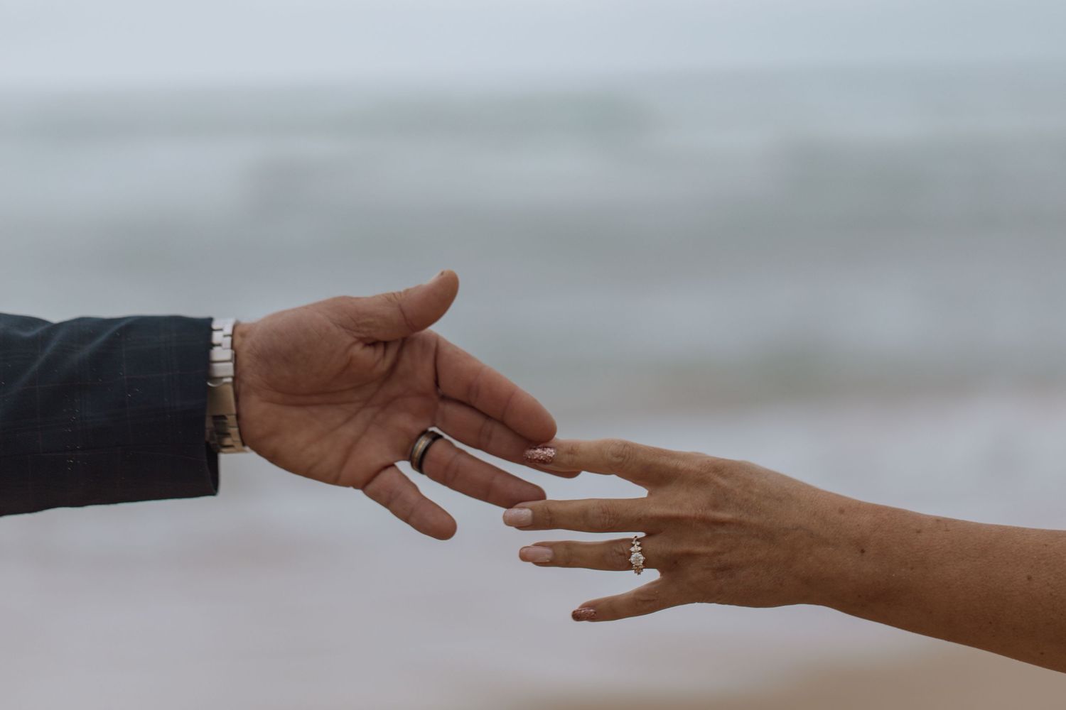 Two hands reach towards each other against a blurry beach background, with one person wearing a dark sleeve.