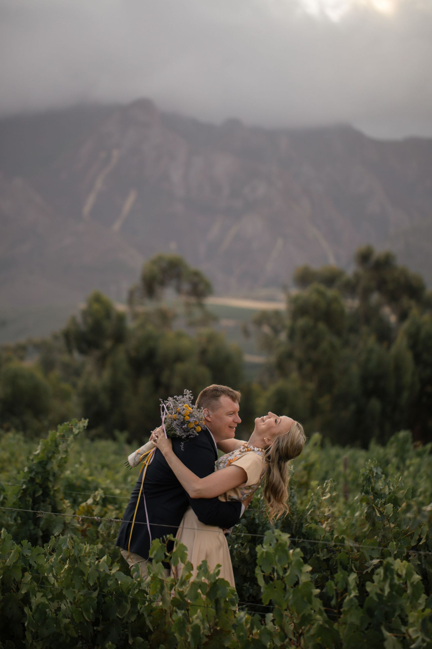 Couple embracing in a vineyard with mountains in the background, man in kilt holding woman with bouquet.