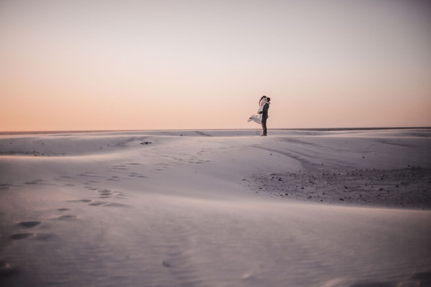 A couple embraces on a vast sandy beach at sunset, silhouetted against a soft pink and orange sky.
