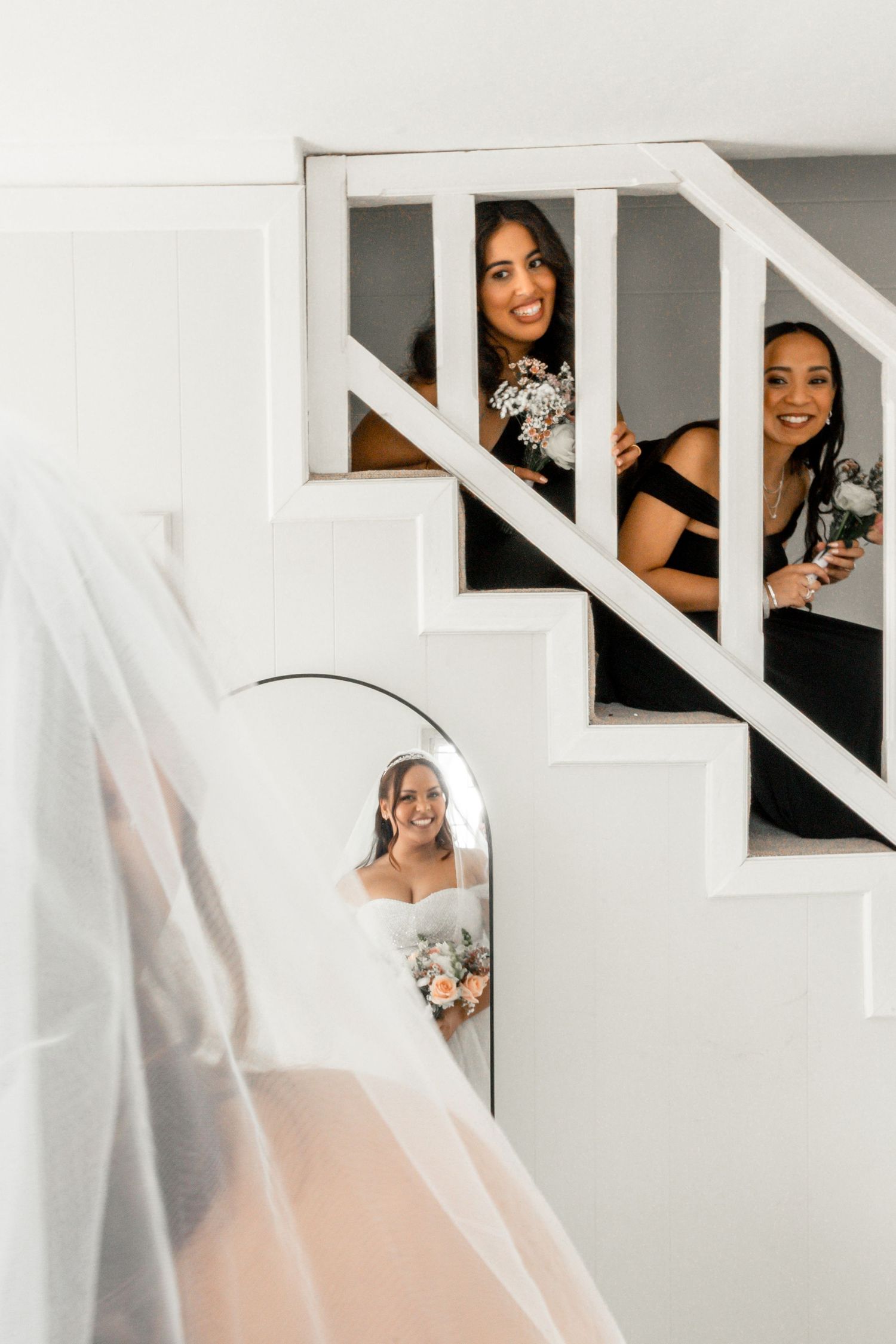 Bride in white gown and veil reflected in mirror while bridesmaids in black dresses peek through staircase railings.