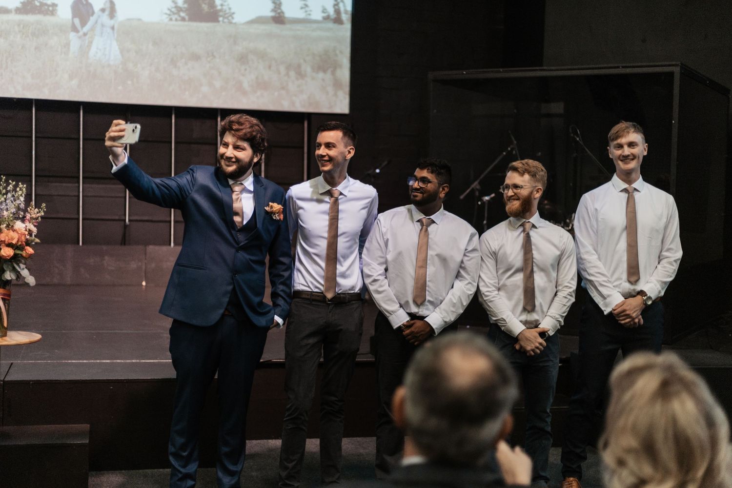 A group of groomsmen in suits and ties stand together on stage at a wedding reception celebration.