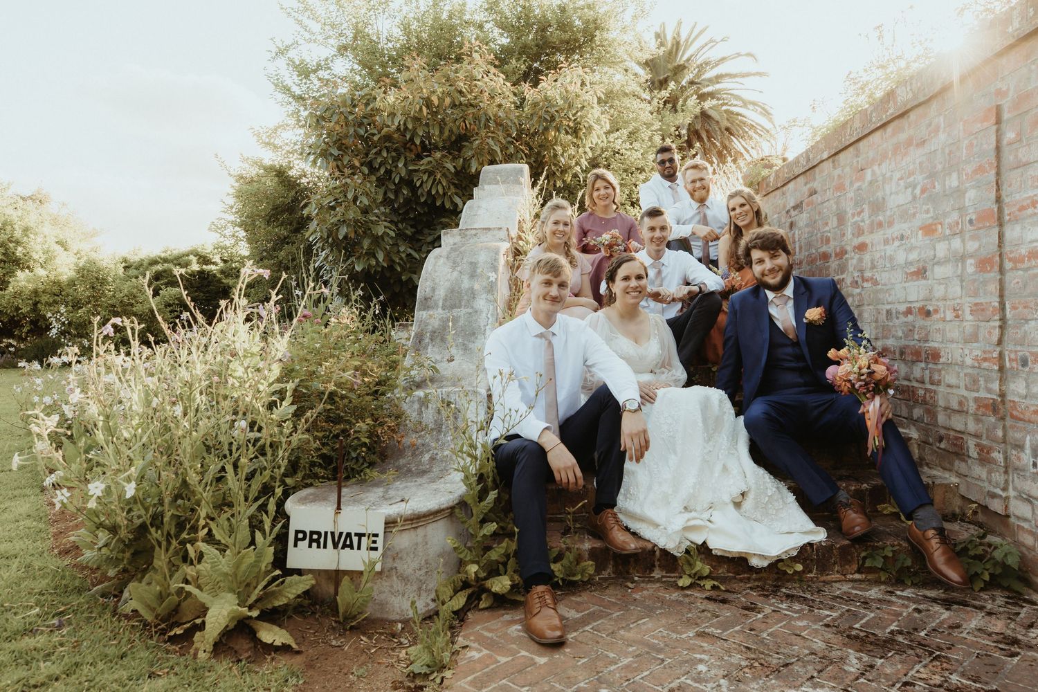 A romantic couple in wedding attire sits together on a rustic stone wall surrounded by lush greenery.