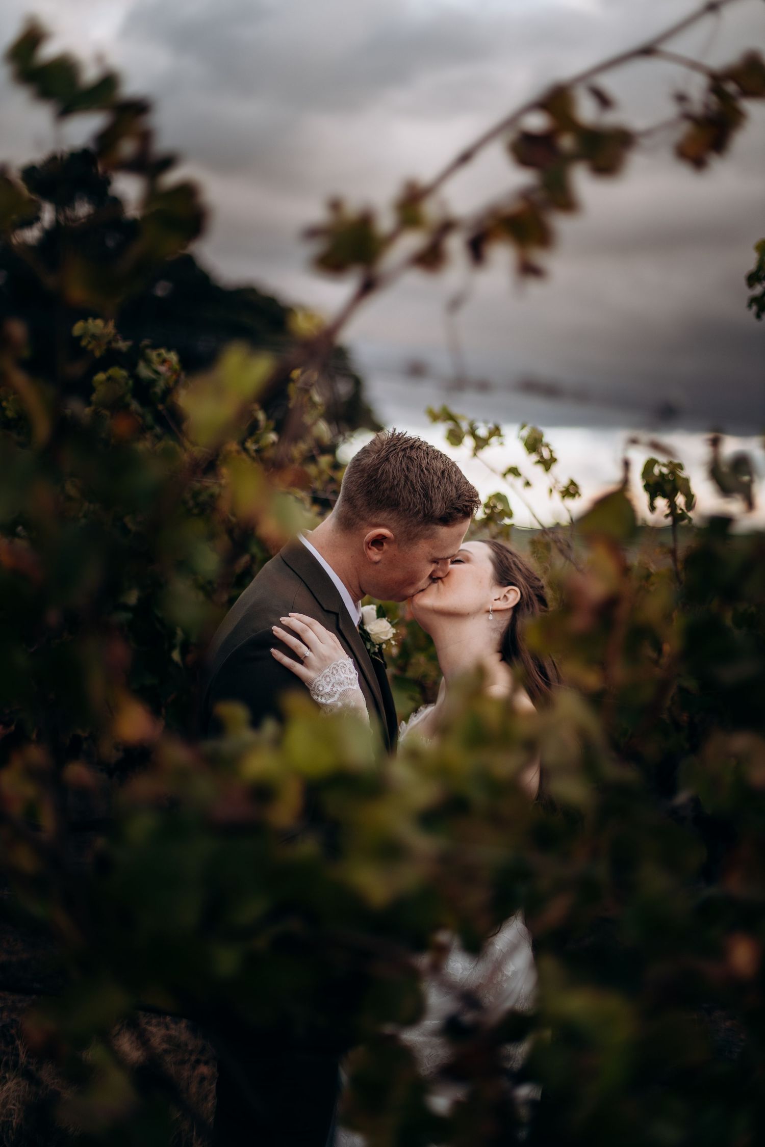 Bride and groom share a romantic kiss surrounded by lush vineyard foliage under a dramatic cloudy sky.