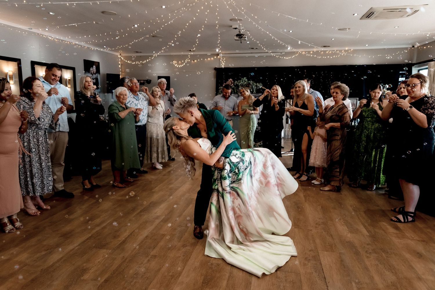 Wedding guests watch a couple dancing together during their first dance at a reception with string lights overhead.