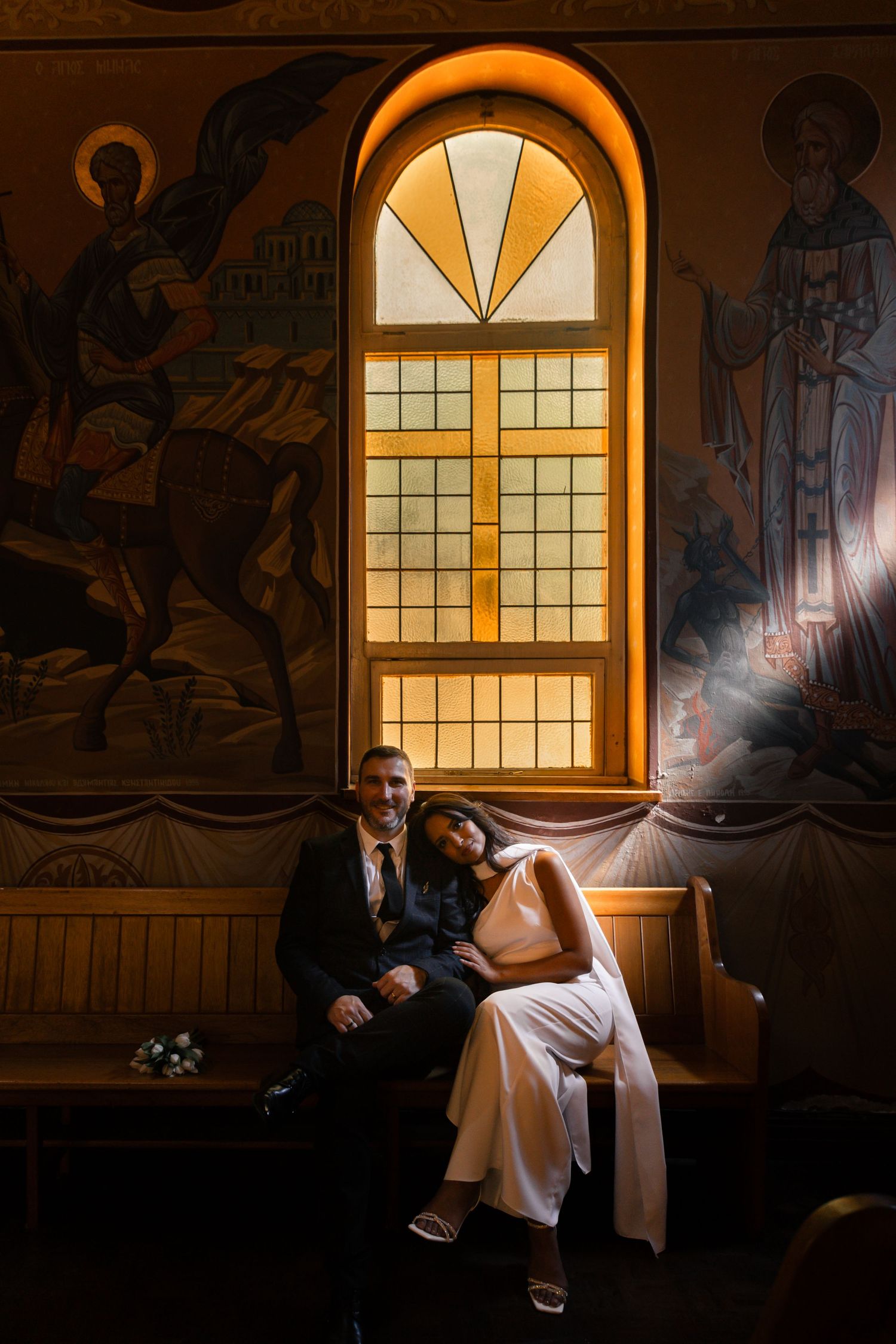 A couple sits together on a wooden pew beneath a dramatic arched window and religious murals in a church interior.
