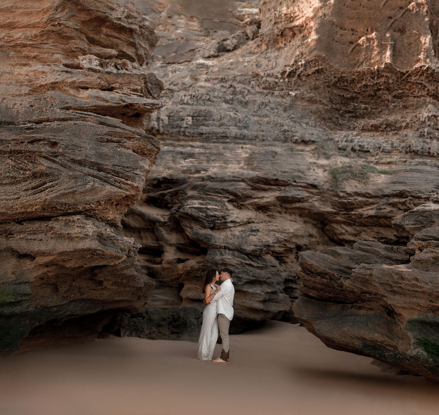 A person in white dress stands beneath a dramatic rocky cave overhang with warm lighting and natural textures.