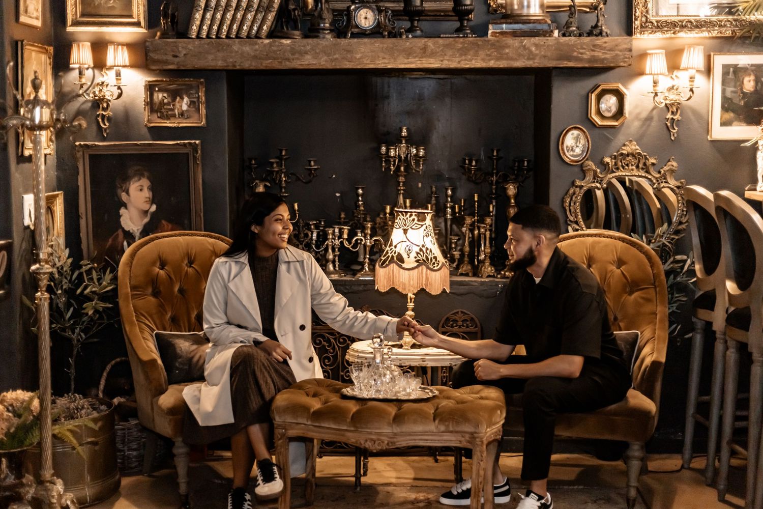 Two people sit in vintage armchairs at a table in a dimly lit antique room with candles and decorative elements.