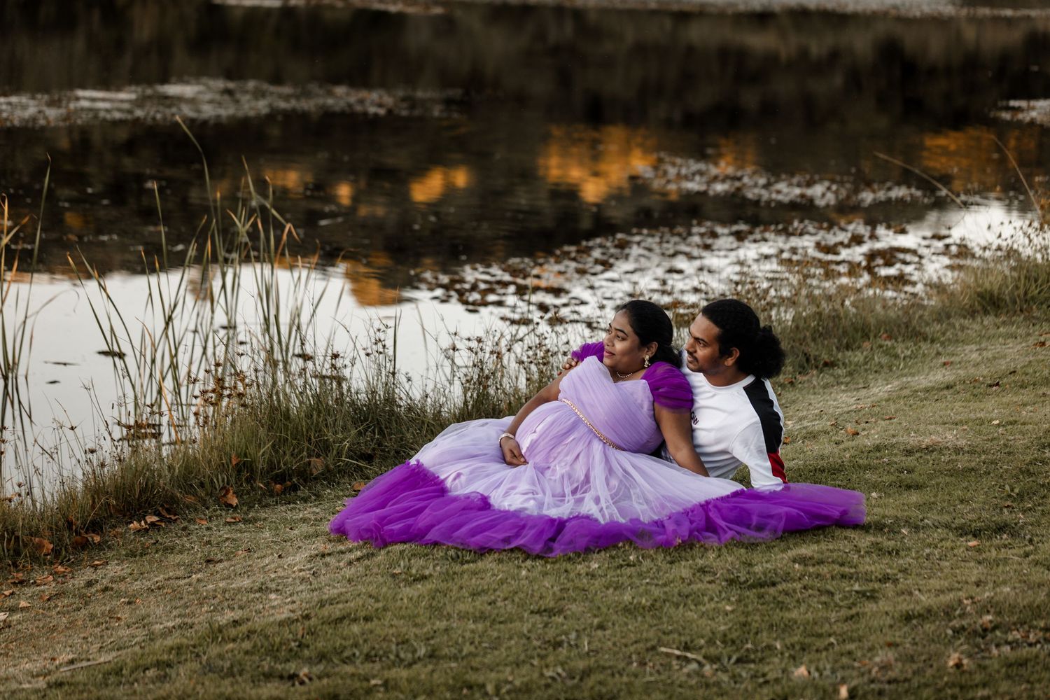 A person in a flowing purple dress lies gracefully on grassy ground during a photo session.