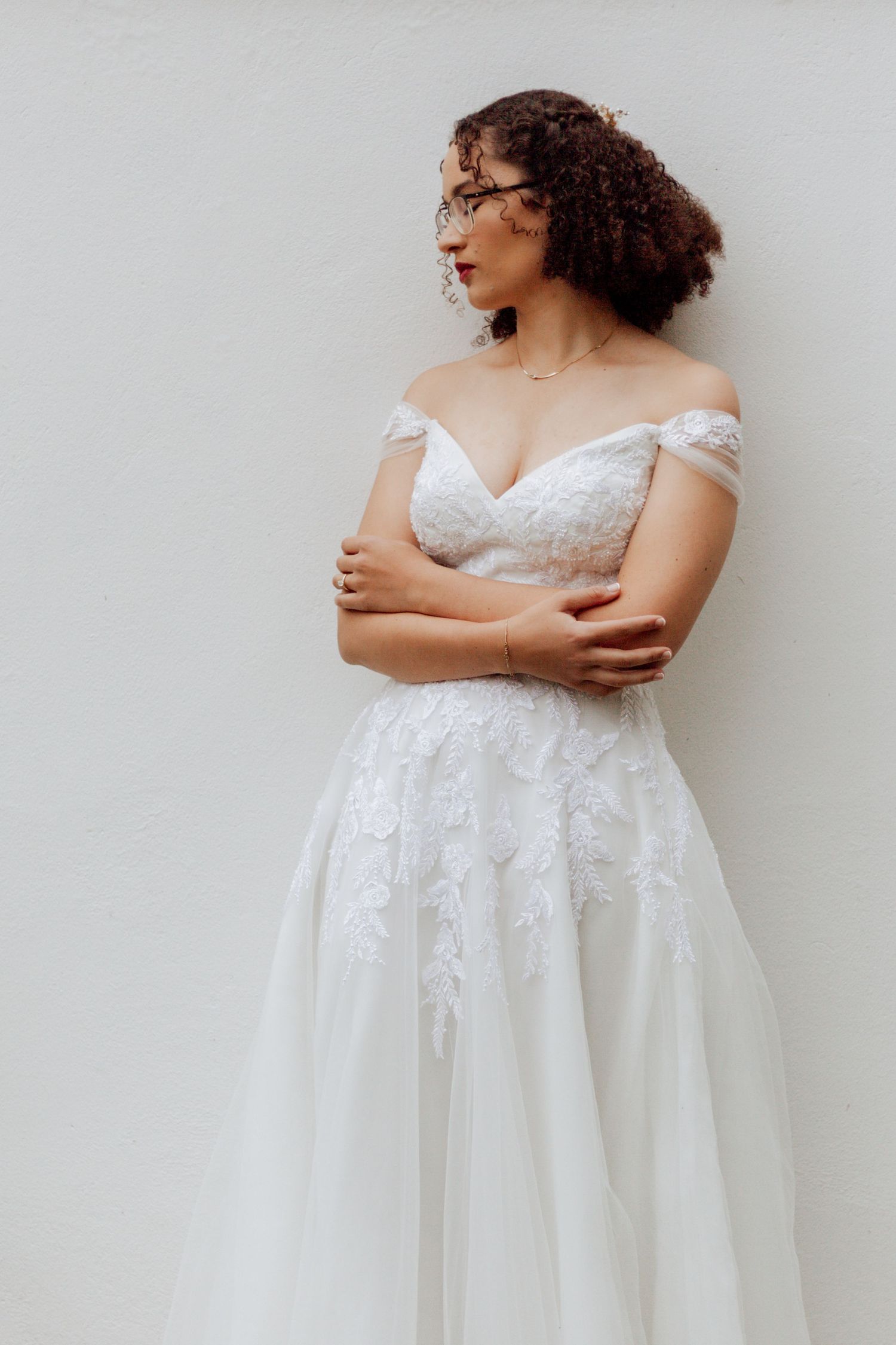 An off-shoulder lace wedding dress with a sweetheart neckline and floral appliques flows elegantly against a white wall.