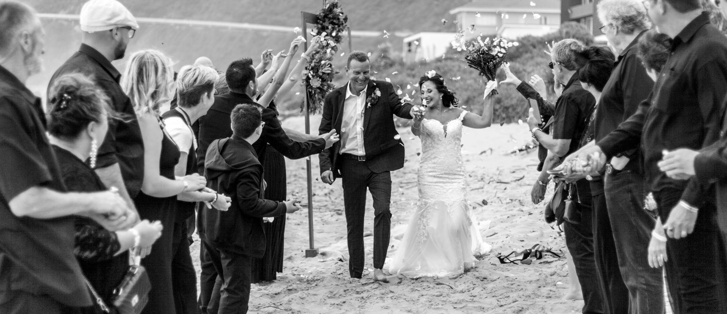 Wedding guests toss flower petals and confetti along a beach path during an outdoor celebration in black and white.