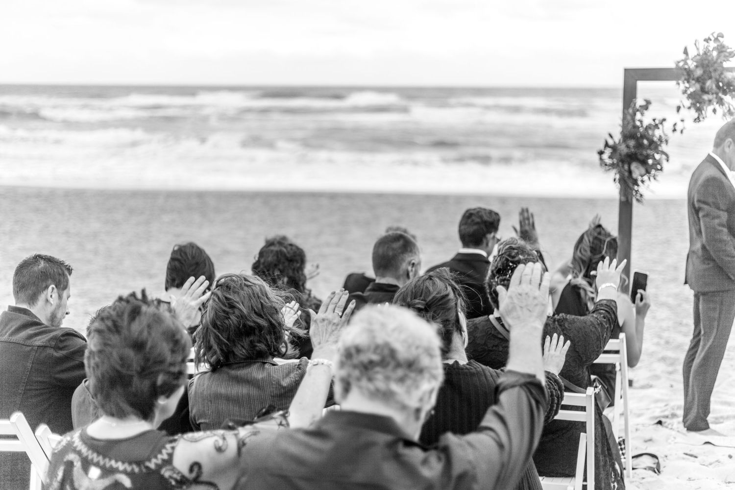 A crowd of spectators watches at beachside seating during a black and white event.