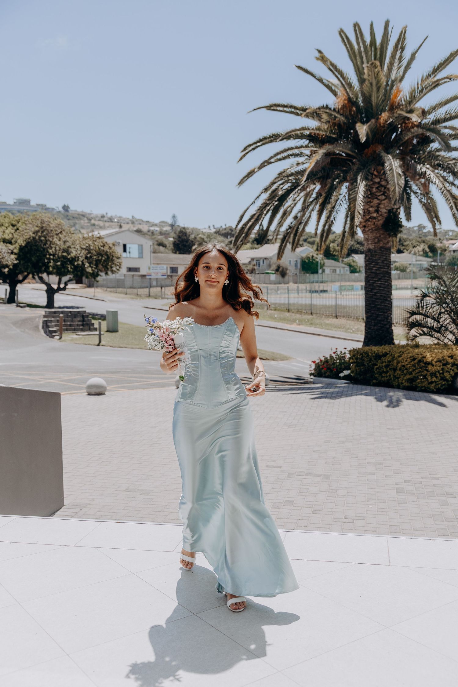 A light blue flowing dress catches the breeze on a sunny day with palm trees and white architecture in the background.