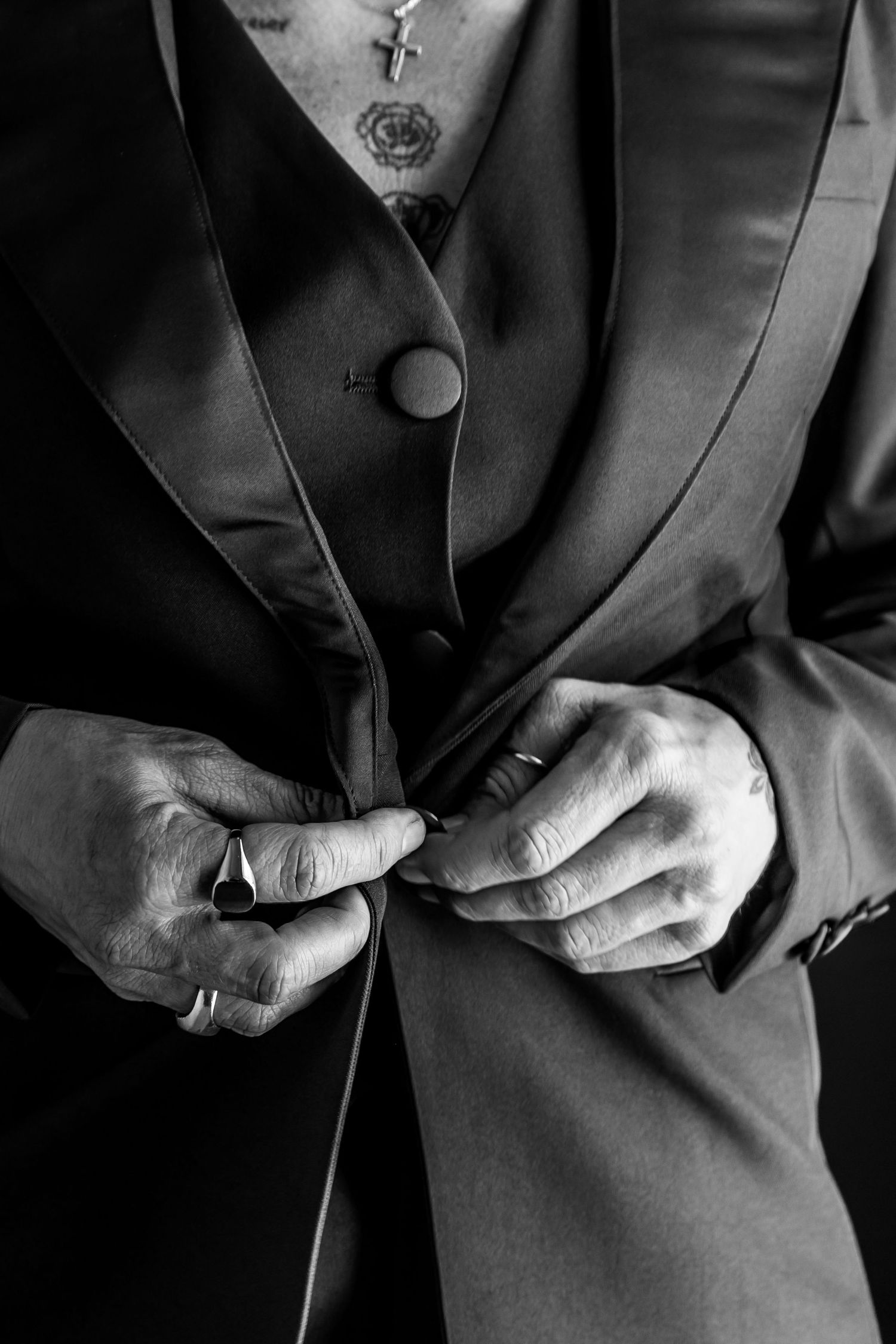 Close-up black and white photo of hands buttoning a formal suit jacket with a visible necklace pendant.