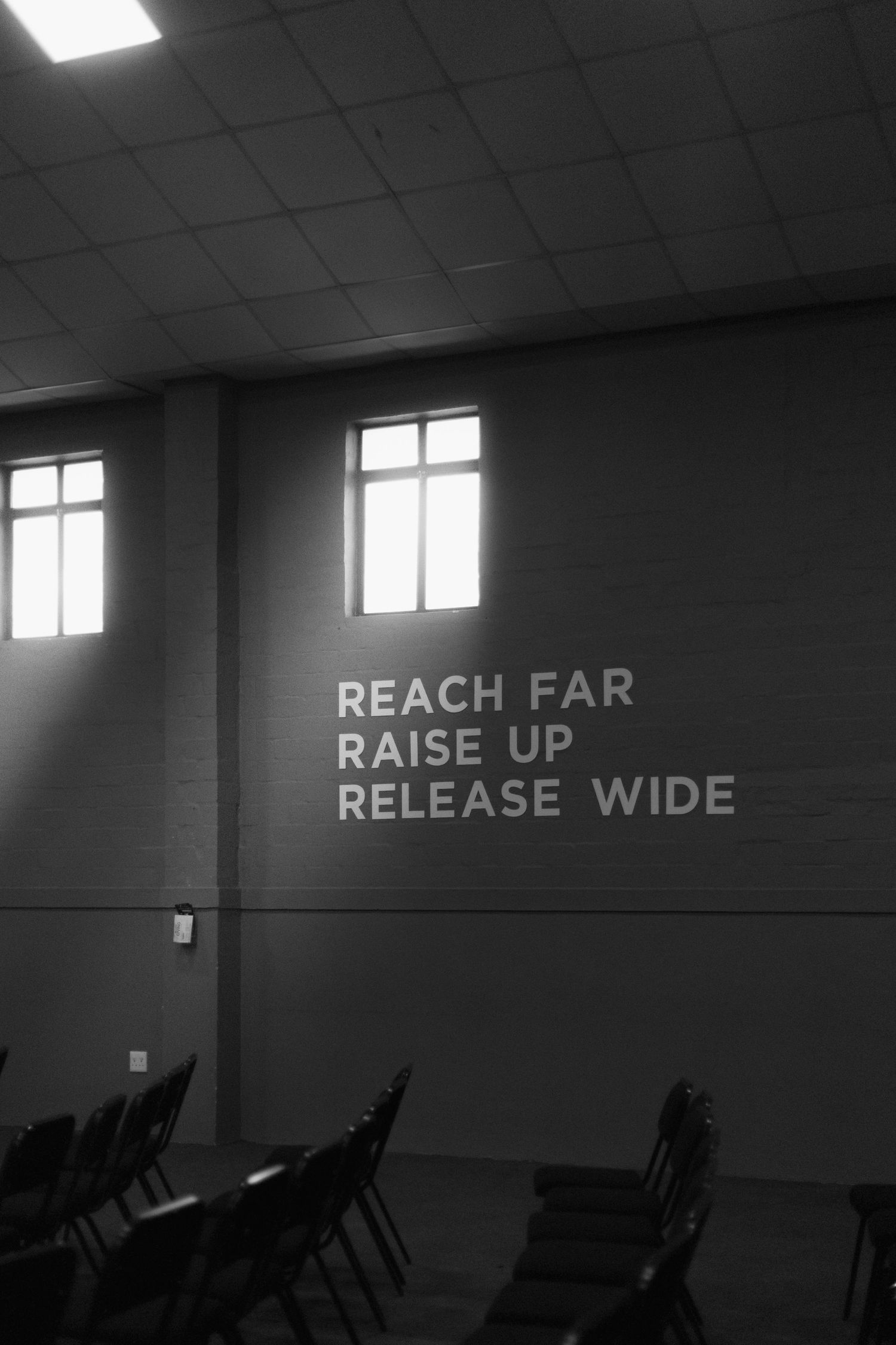 Black and white photo of an empty room with folding chairs and text on wall reading 'REACH FAR RAISE UP RELEASE WIDE'.