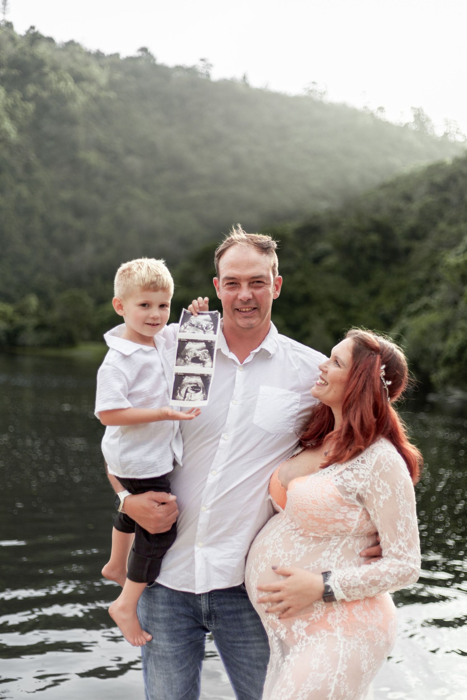 Family poses by a lake surrounded by lush hills while holding ultrasound photos and showing a pregnant belly.