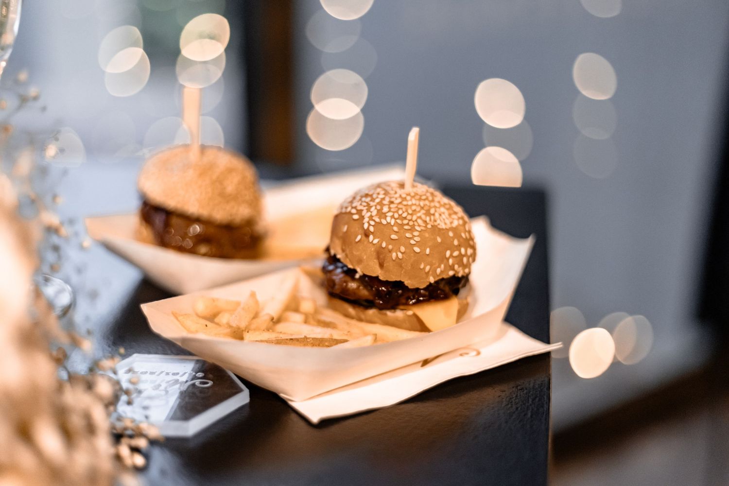 A gourmet burger with sesame seed bun and french fries served on wooden board with sparkly bokeh background.