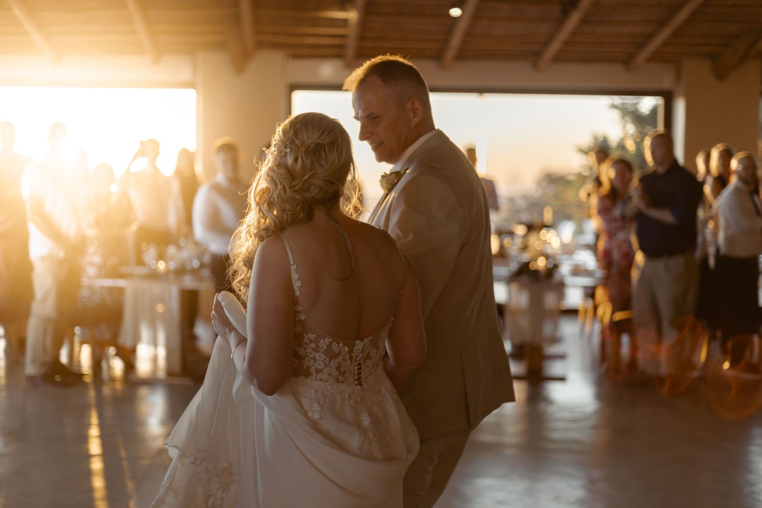 A newlywed couple shares their first dance at sunset during their wedding reception with guests in the background.