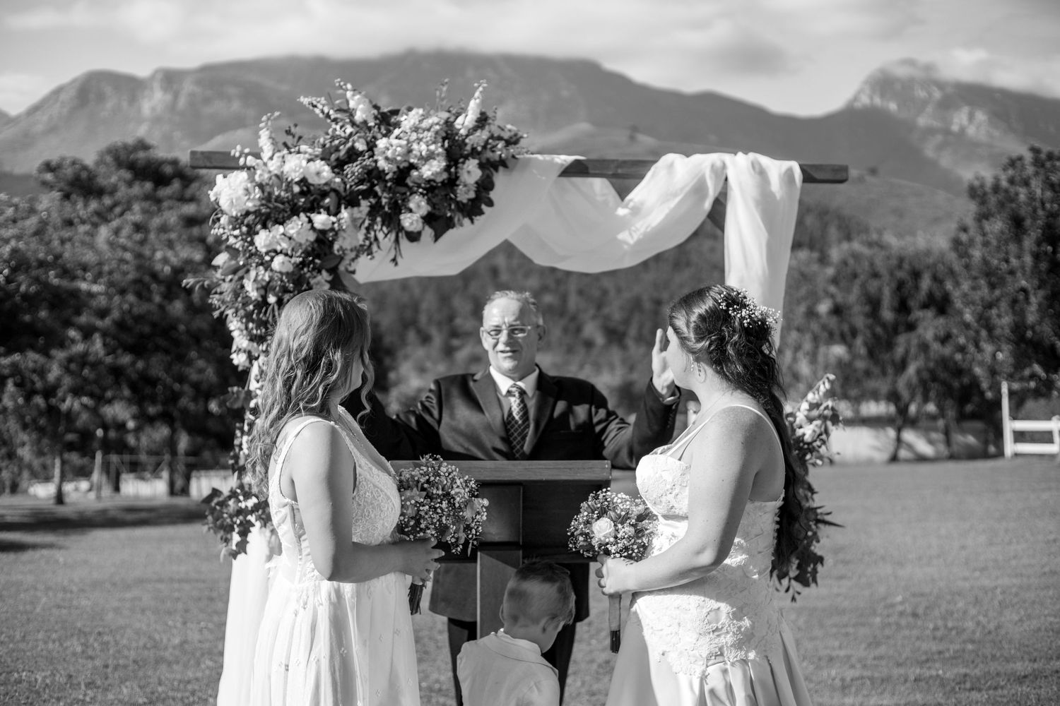 An outdoor wedding ceremony with a draped arch and floral arrangements takes place against a backdrop of rolling mountains.