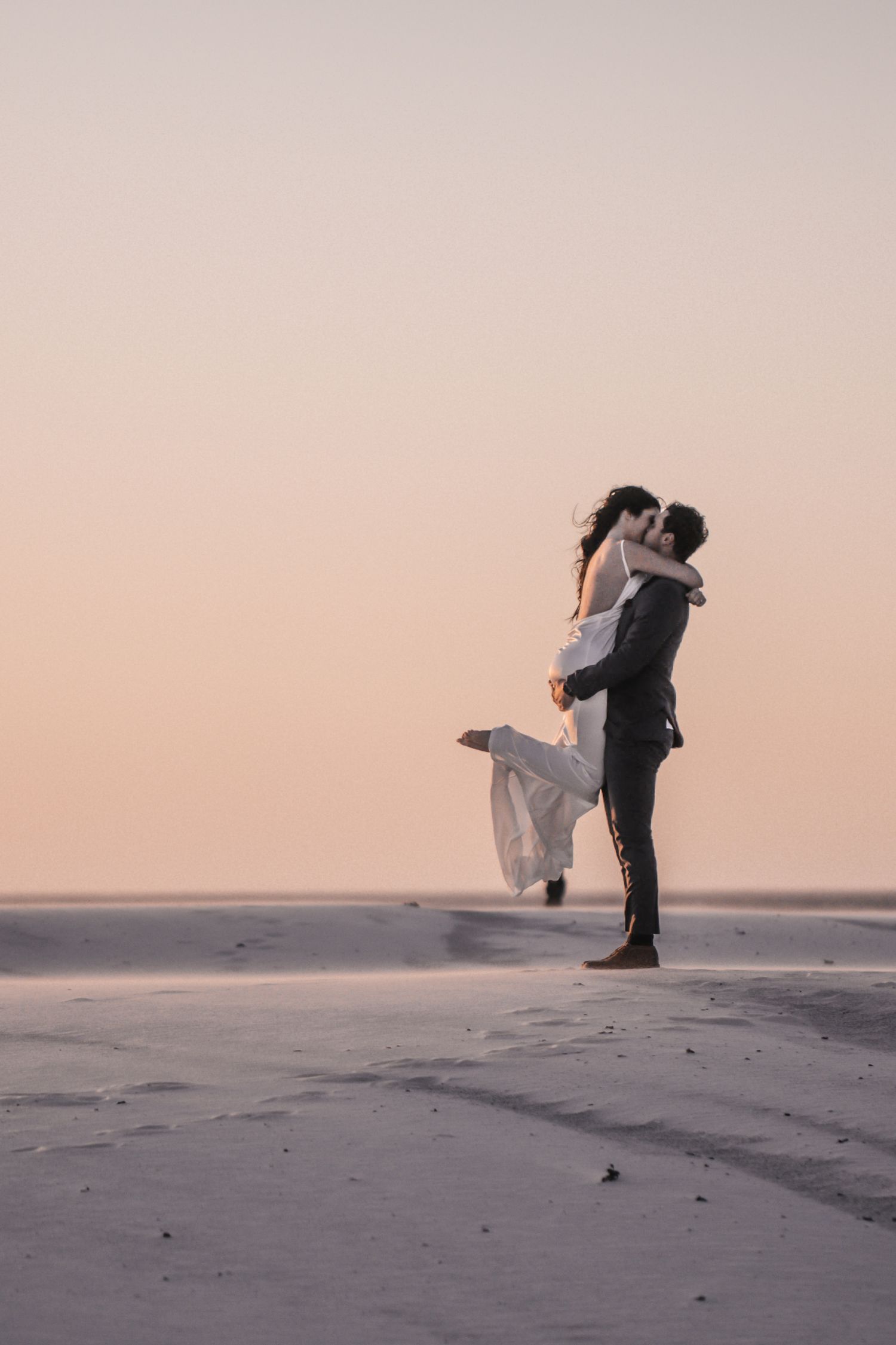 A groom lifts his bride on a sandy beach at sunset, sharing a romantic kiss in a dreamy golden light.