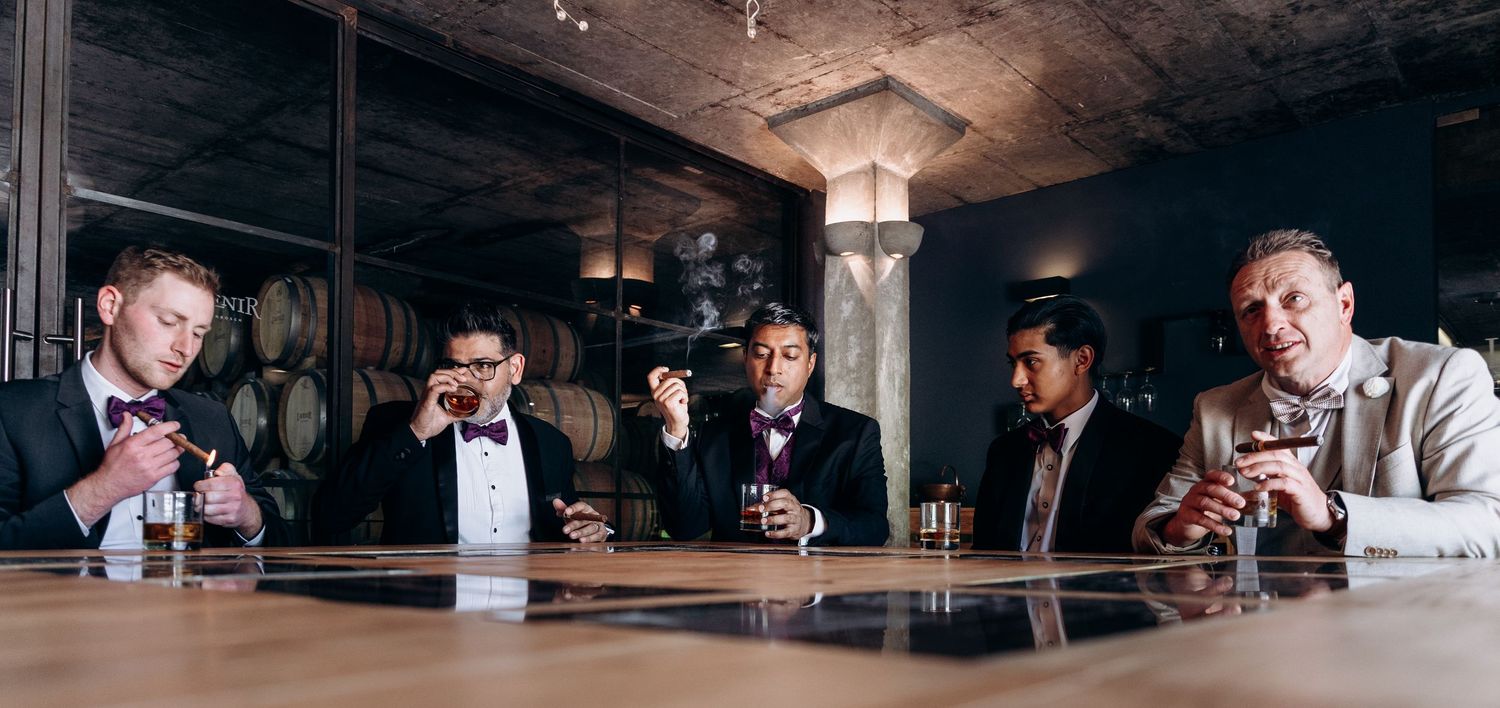 Five well-dressed men in suits enjoying drinks and cigars at a stylish bar with wine barrels in the background.