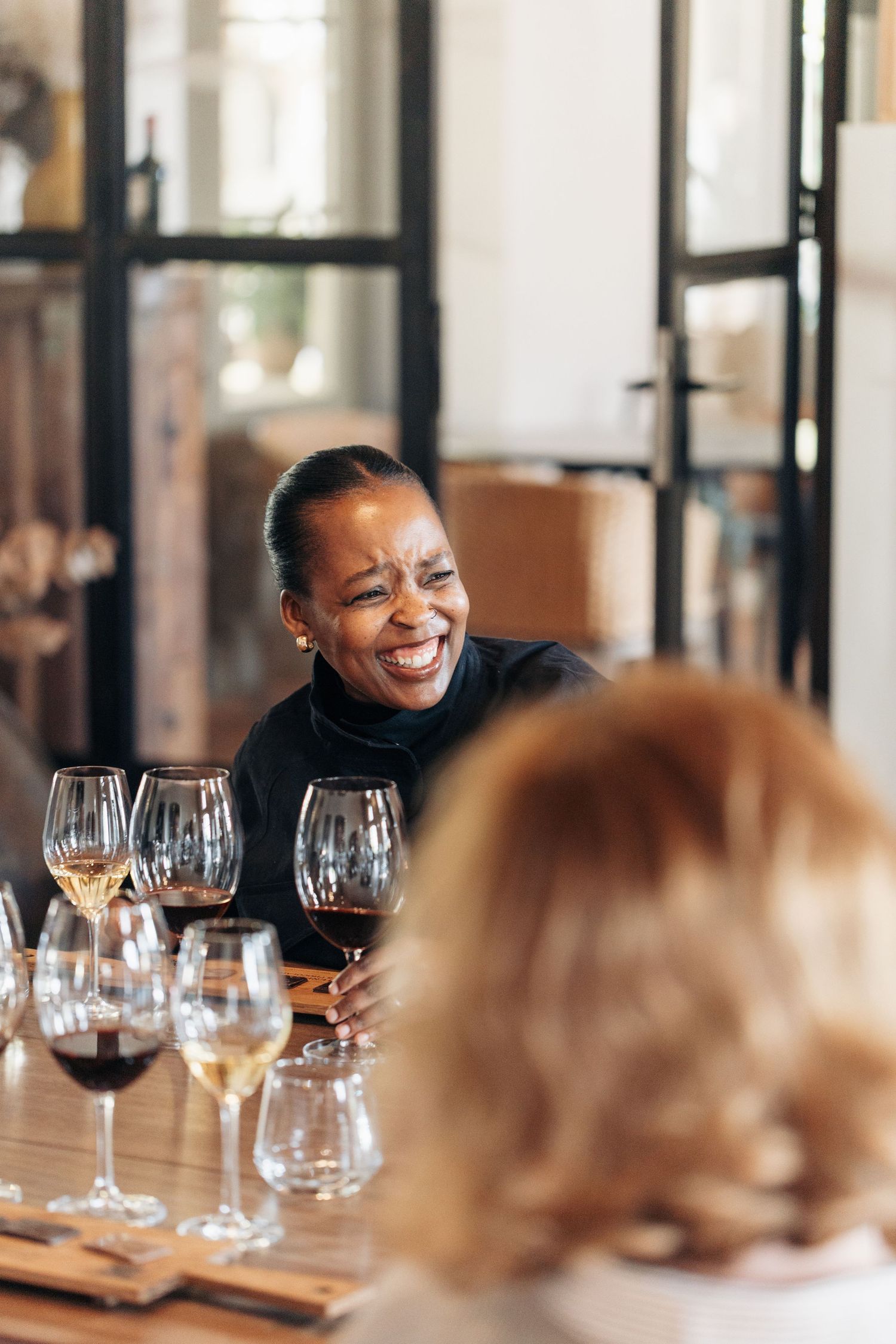 A smiling woman enjoys a wine tasting session at a wooden table filled with various glasses of red and white wine.