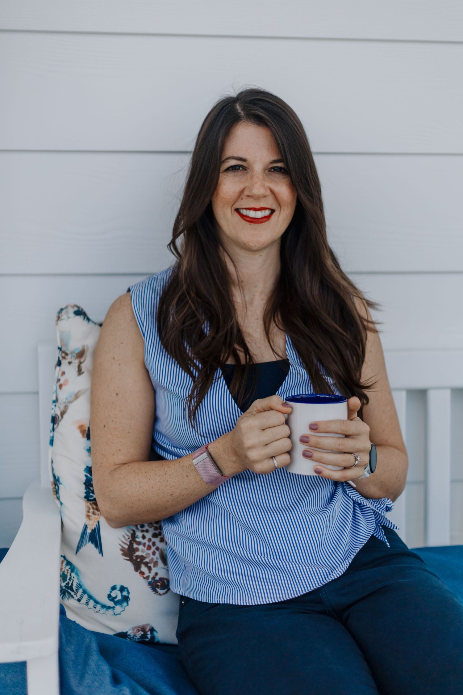 A woman in a blue sleeveless top sits on a porch with a coffee mug and floral tote bag.