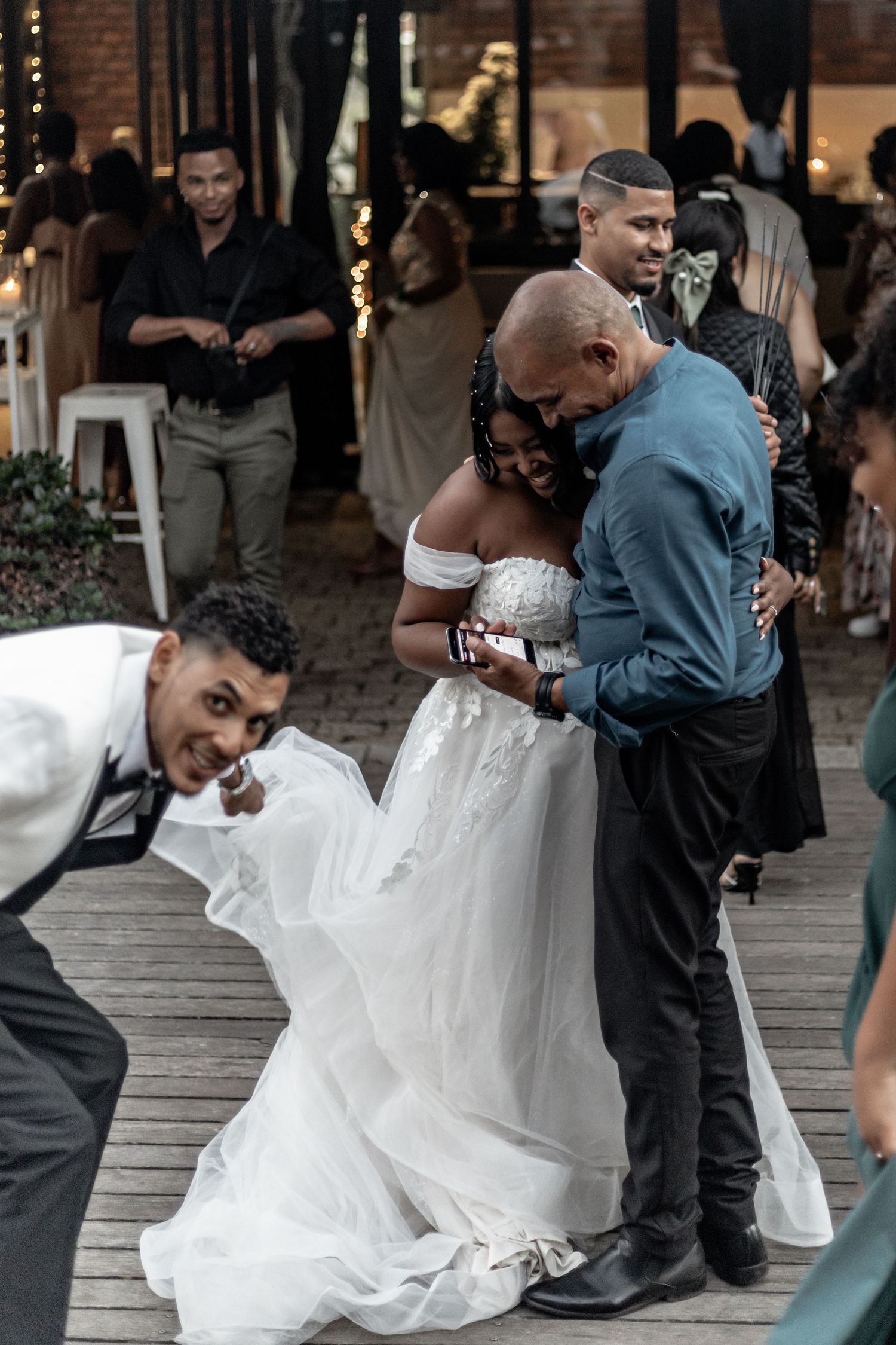 A candid moment during a wedding reception shows a guest helping adjust the bride's flowing white dress on a wooden deck.