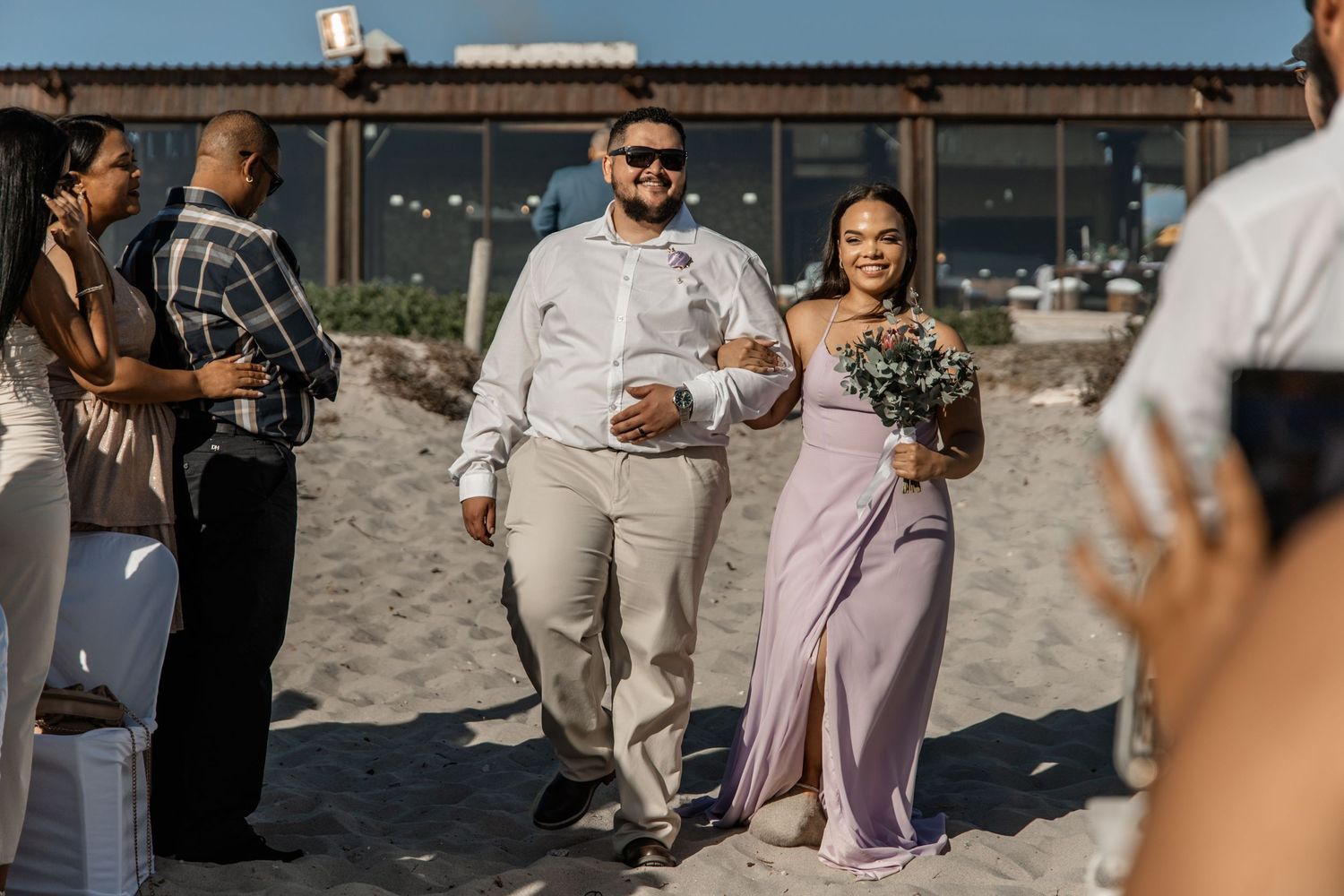 Wedding party celebrates on a sunny beach with guests in beige and pink attire gathering by the ocean.