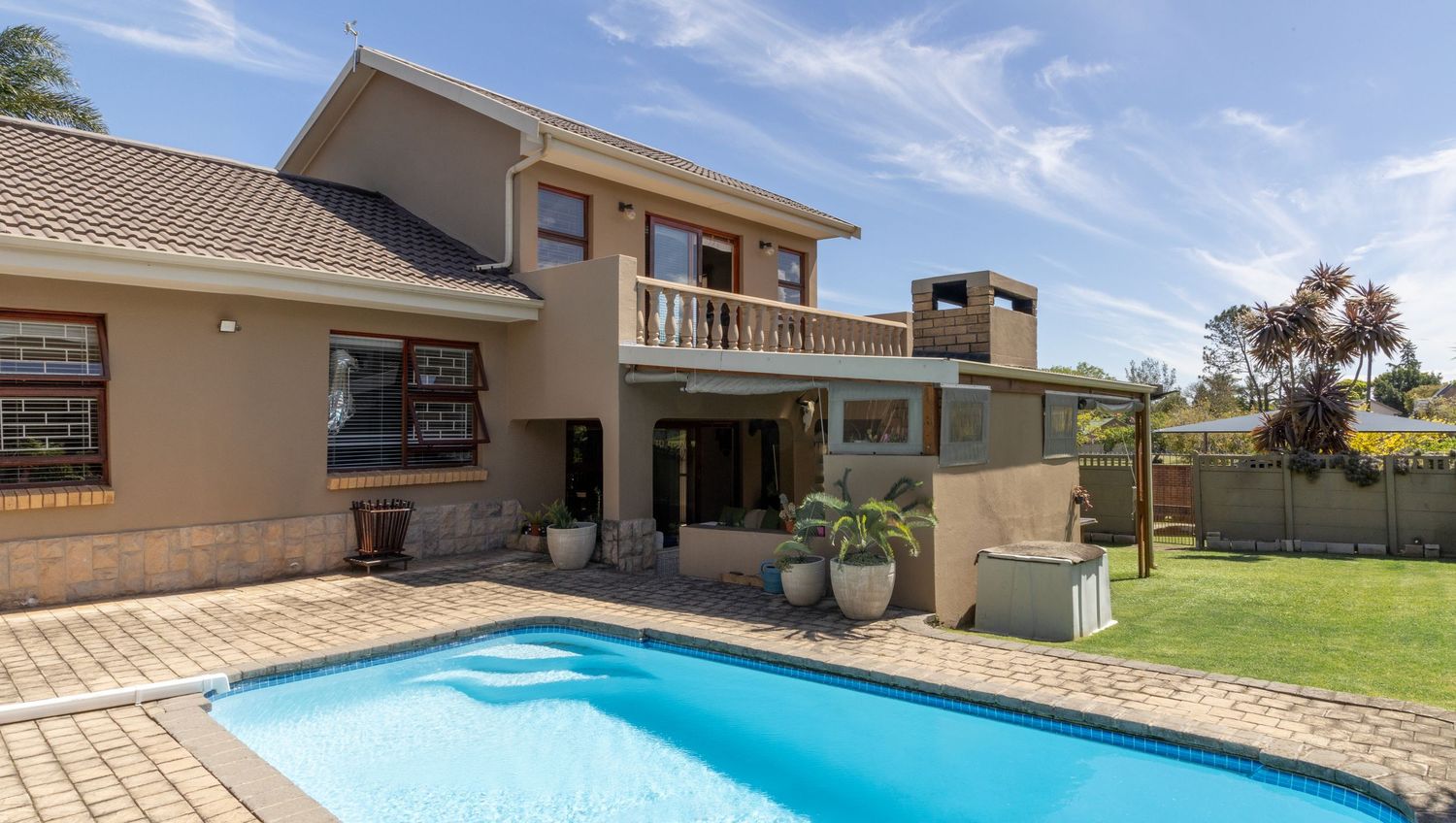 Modern beige two-story house with balcony and blue swimming pool in backyard under sunny sky.