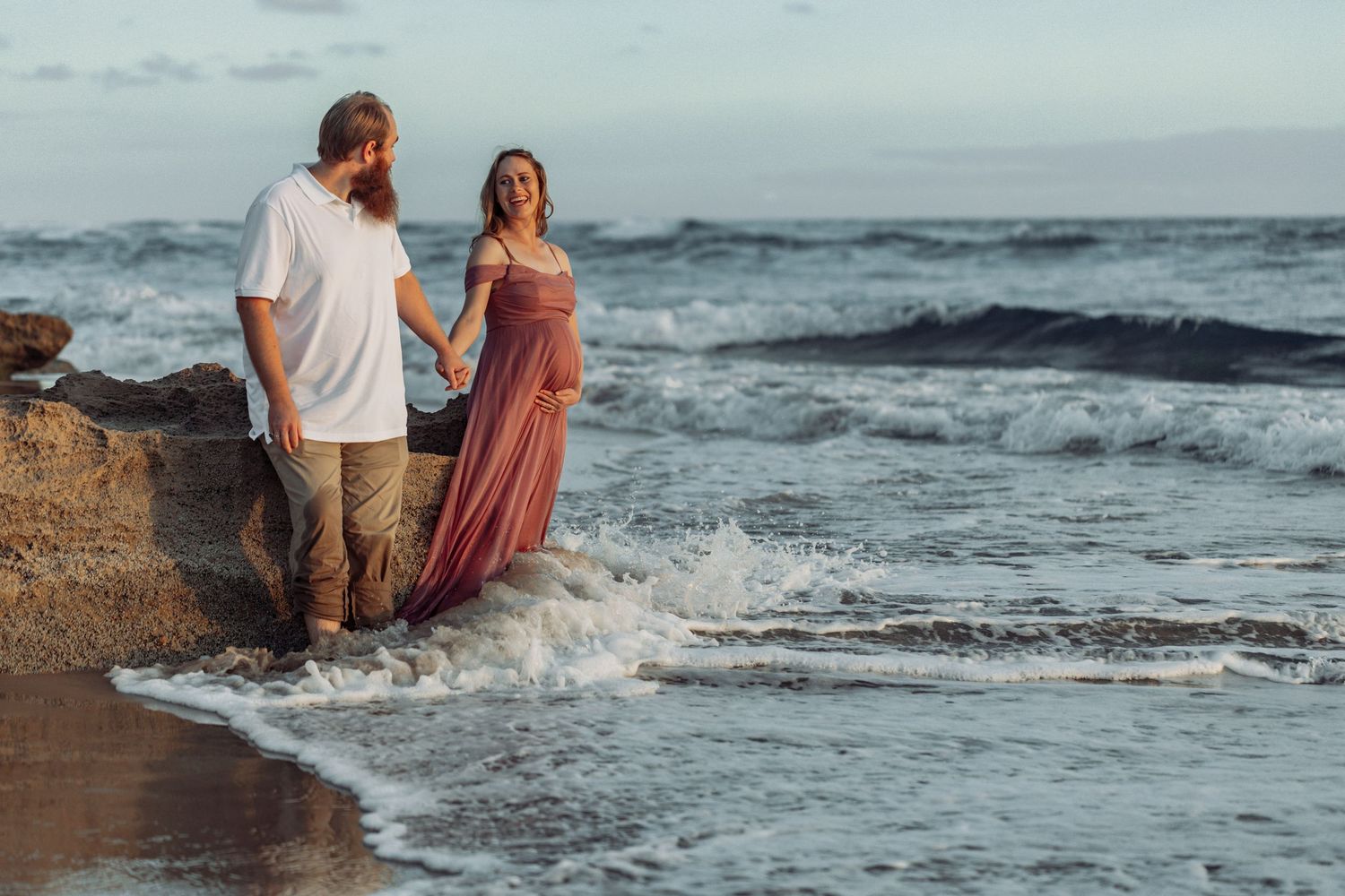 Couple walking along the beach shore with waves crashing and sunset in the background.