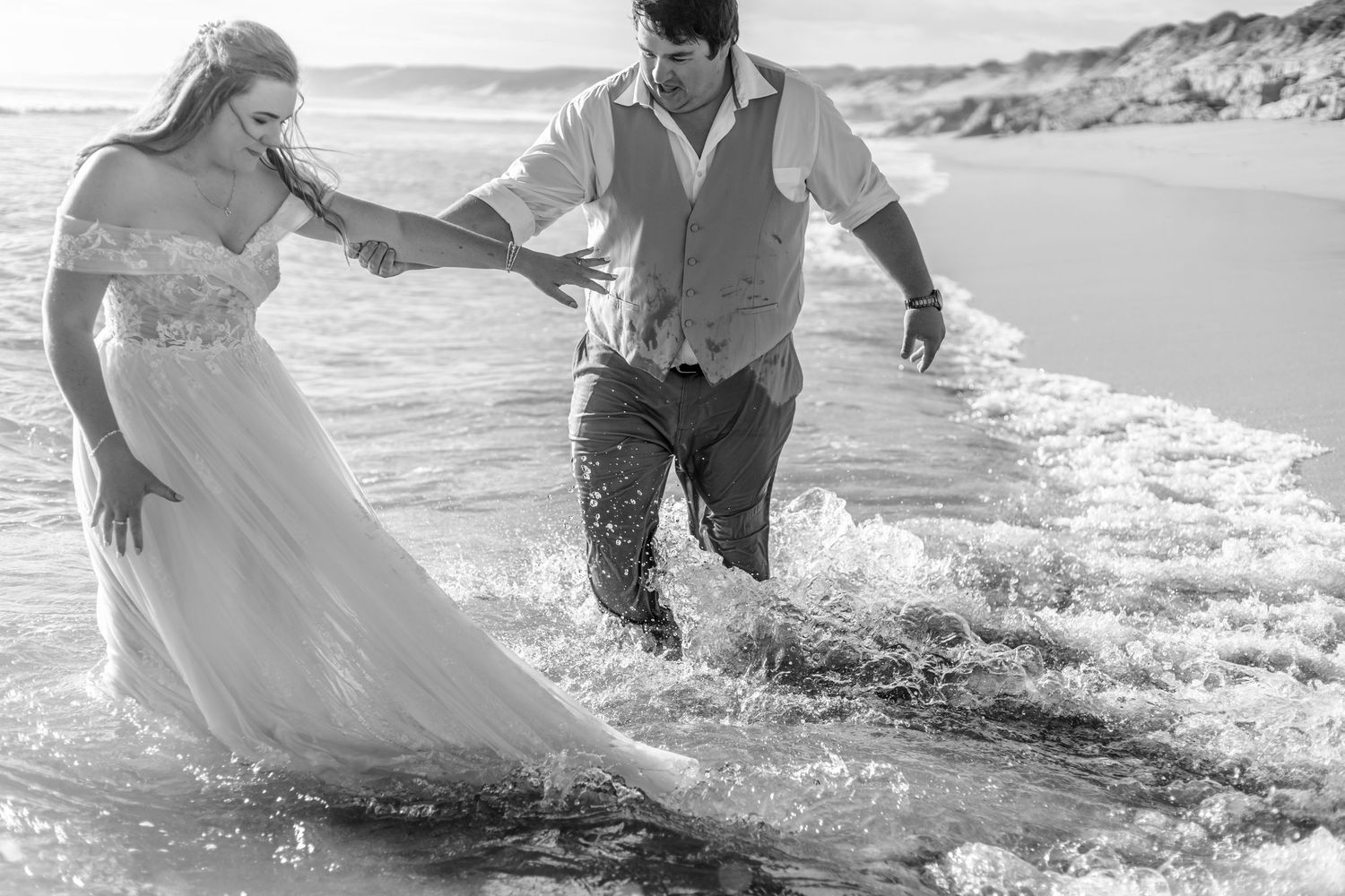 A couple in formal attire runs playfully through ocean waves at the beach during a romantic photoshoot.