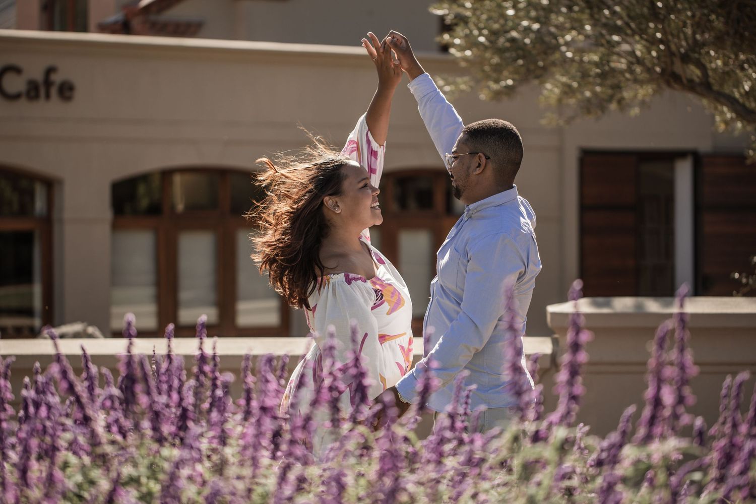 A couple walks hand in hand through a garden with blooming purple flowers on a sunny day.