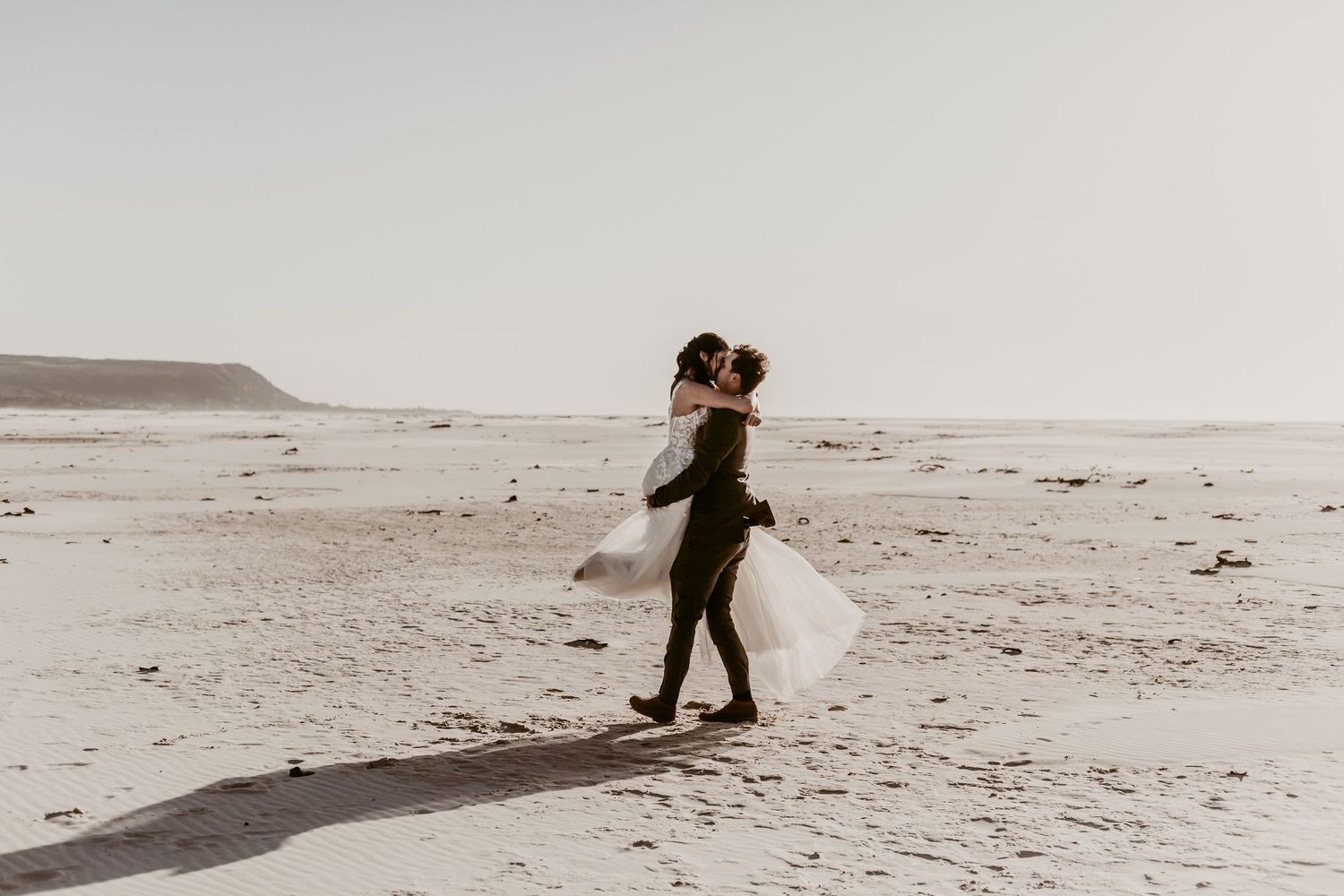 A couple shares a romantic embrace on a sandy beach, the bride in a flowing white gown lifted by her partner.