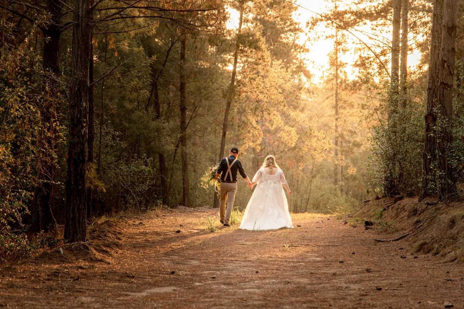 Bride and groom walking hand in hand down a sunlit forest path surrounded by autumn trees.