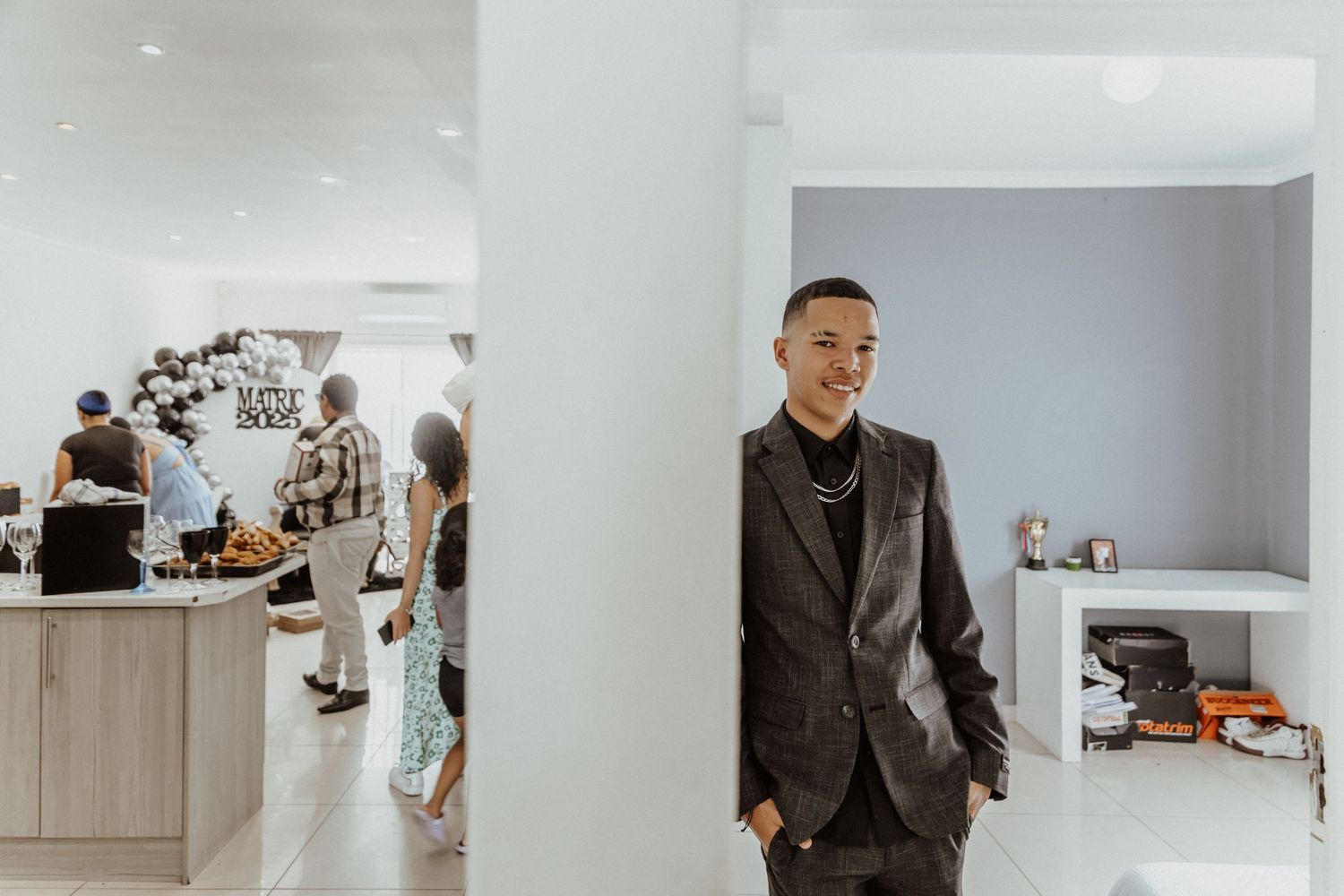 Young man in dark suit smiling at a modern white interior event space with balloon decorations.