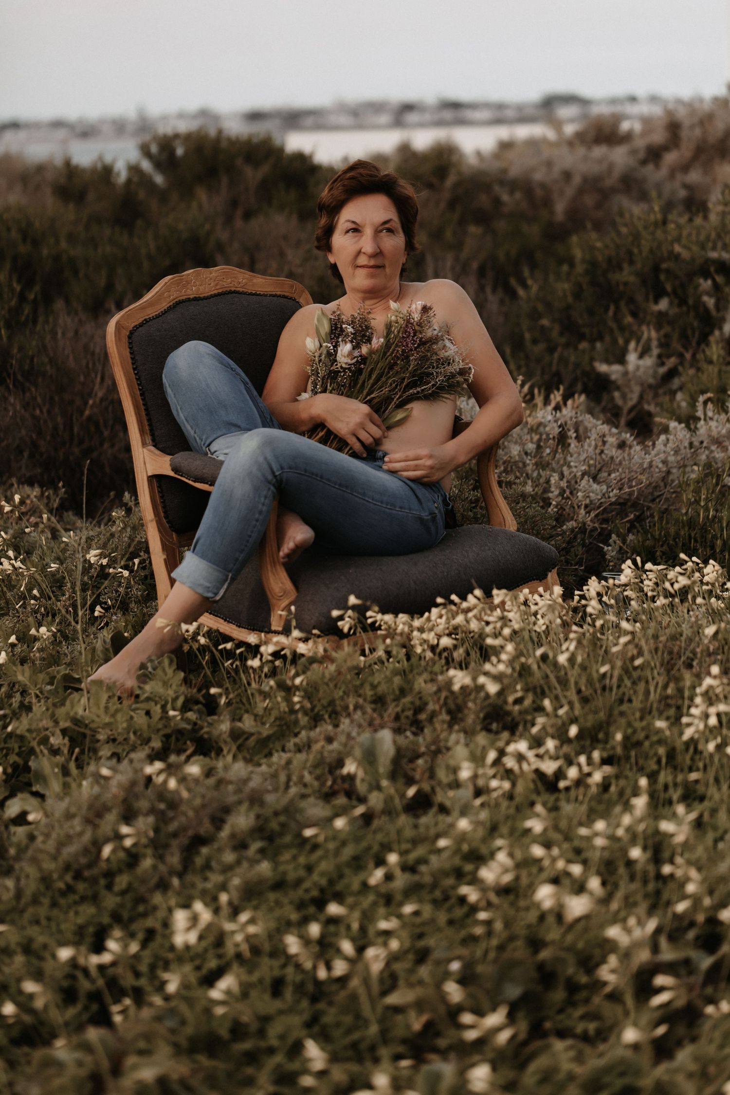 A person sits barefoot in a vintage chair among wildflowers while holding dried flowers at sunset.