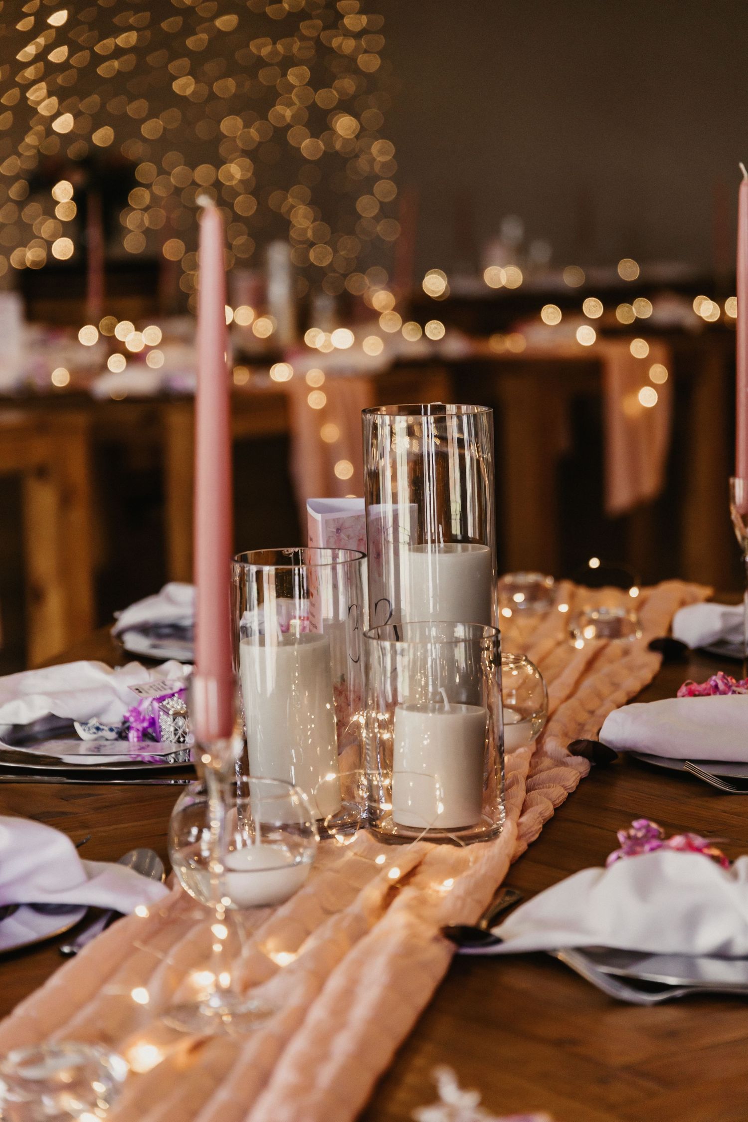 Elegant wedding reception table setting with pink candles, white flowers and glowing bokeh lights in the background.