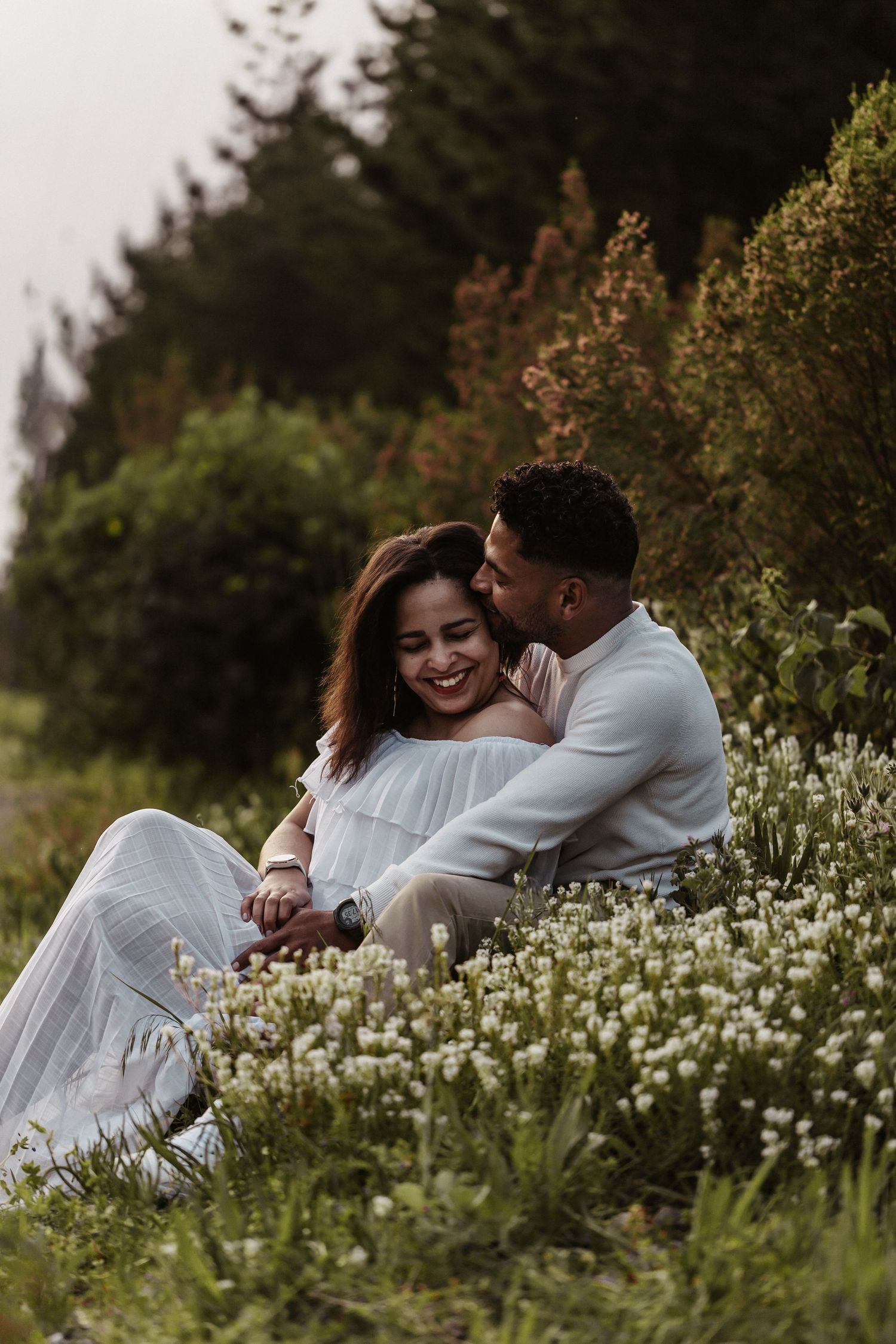 A couple in white clothing sits intimately together among blooming white wildflowers with evergreen trees in background.