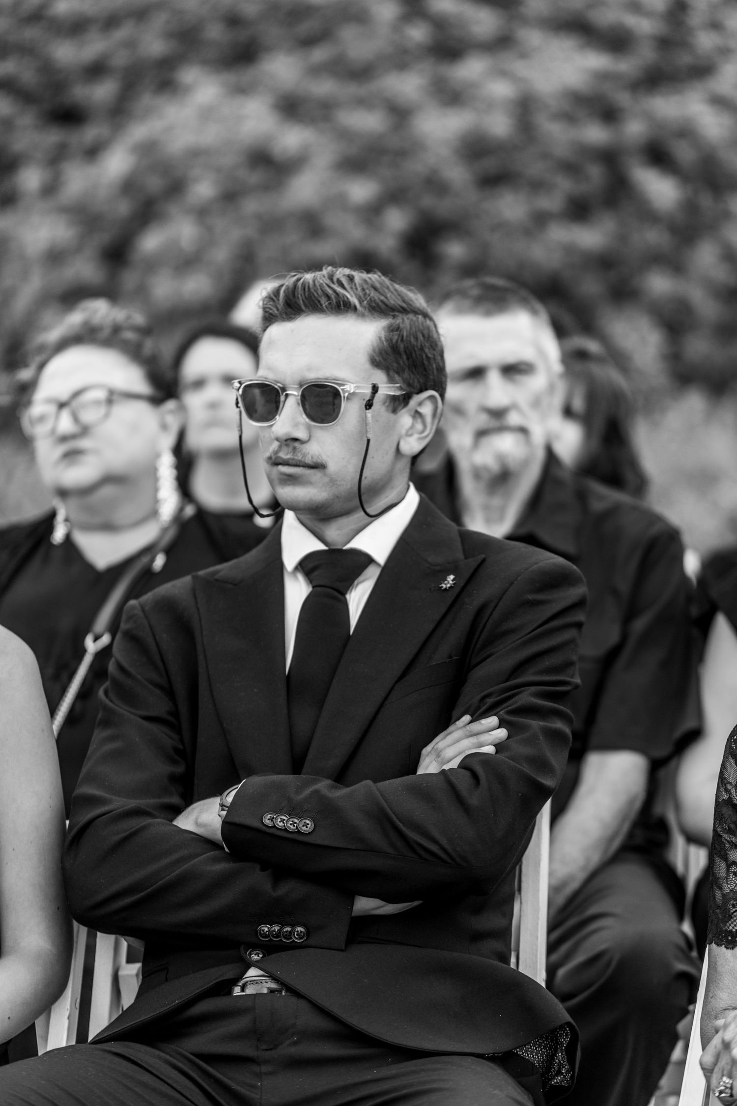 Black and white photo of someone in sunglasses and suit sitting with arms crossed at formal event with others in background.