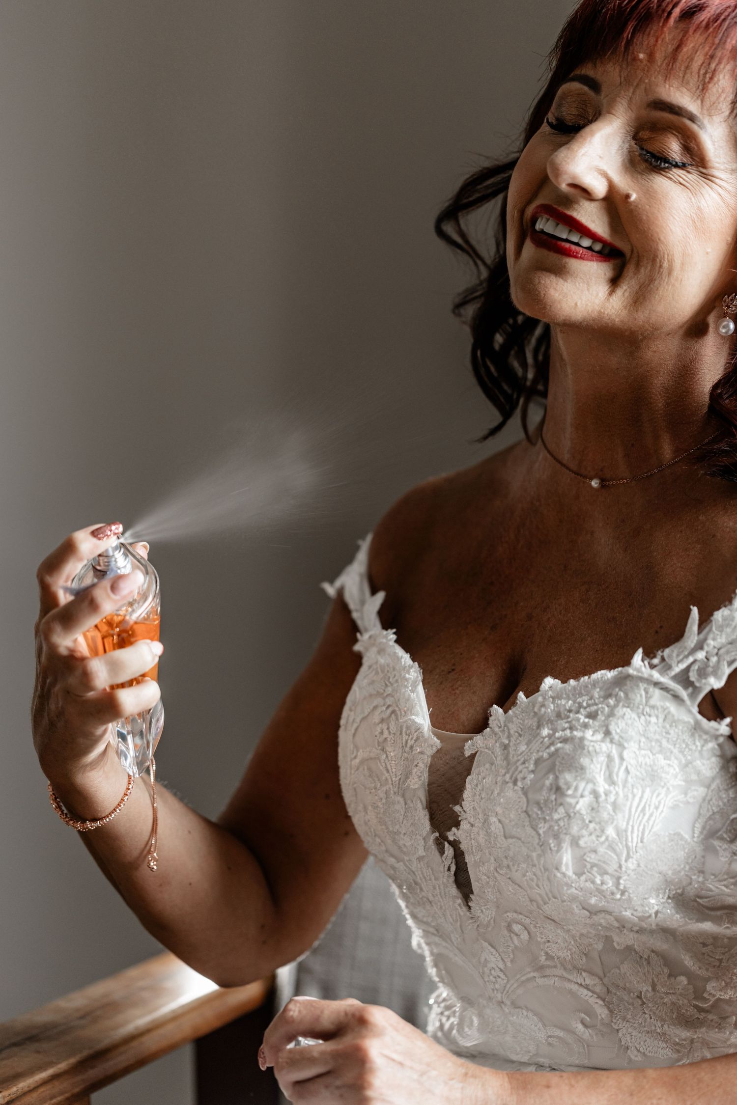 A smiling person in a white lace wedding dress sprays perfume in a softly lit setting.
