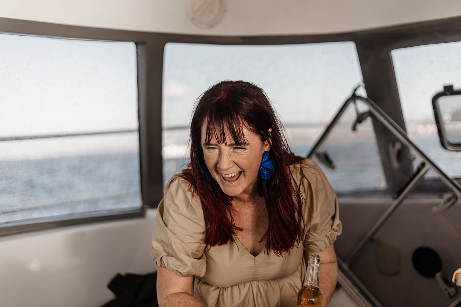 A person with blue earrings laughing joyfully inside a boat cabin with ocean views.