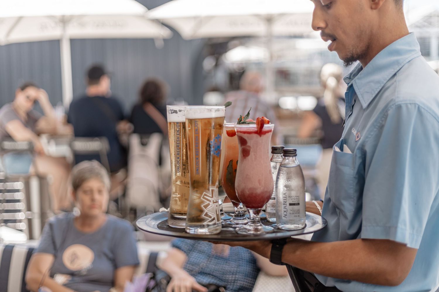 Server carries a tray with tall glasses of beer and a red cocktail at an outdoor restaurant patio.