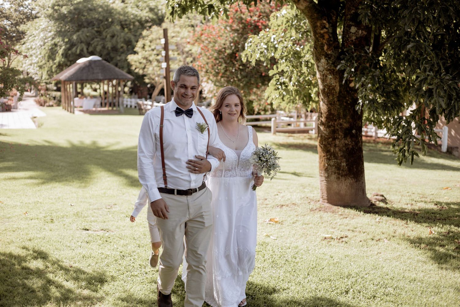 A couple walks arm in arm through a sunny garden setting during their outdoor wedding celebration.