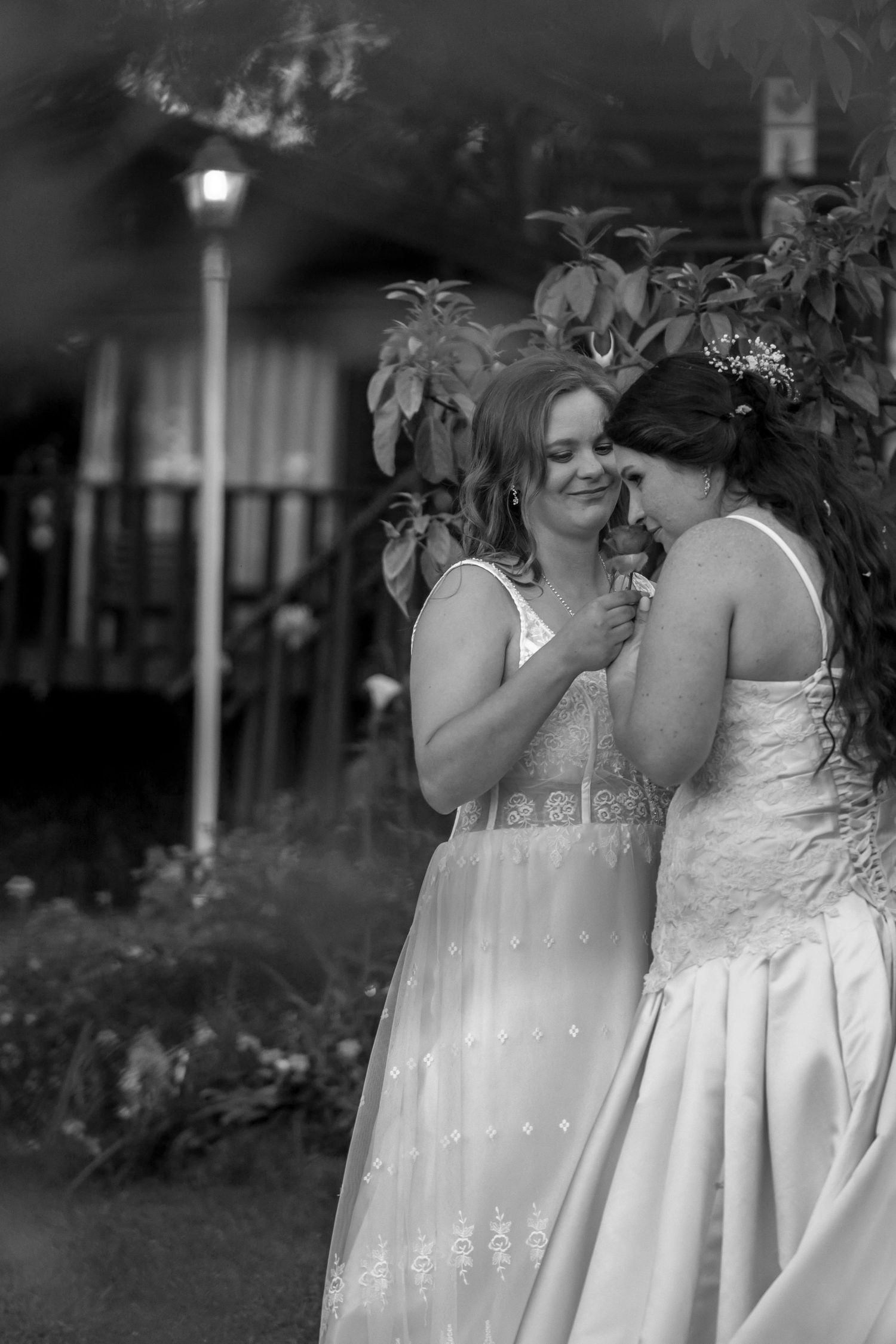 Two people in elegant white dresses share an intimate moment in a garden setting at dusk, captured in black and white.