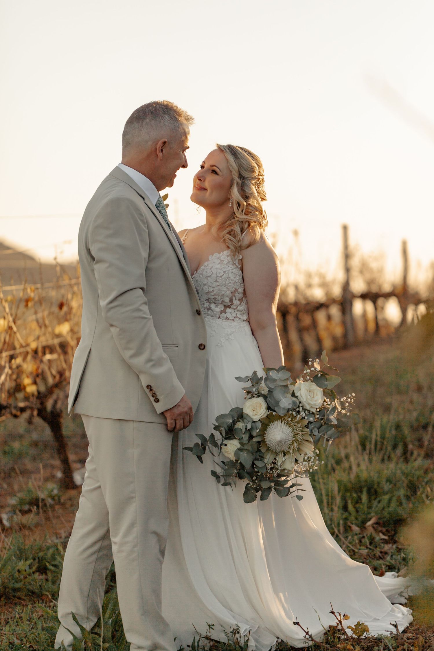 A bride and groom share an intimate moment in a vineyard at sunset while she holds a white floral bouquet.