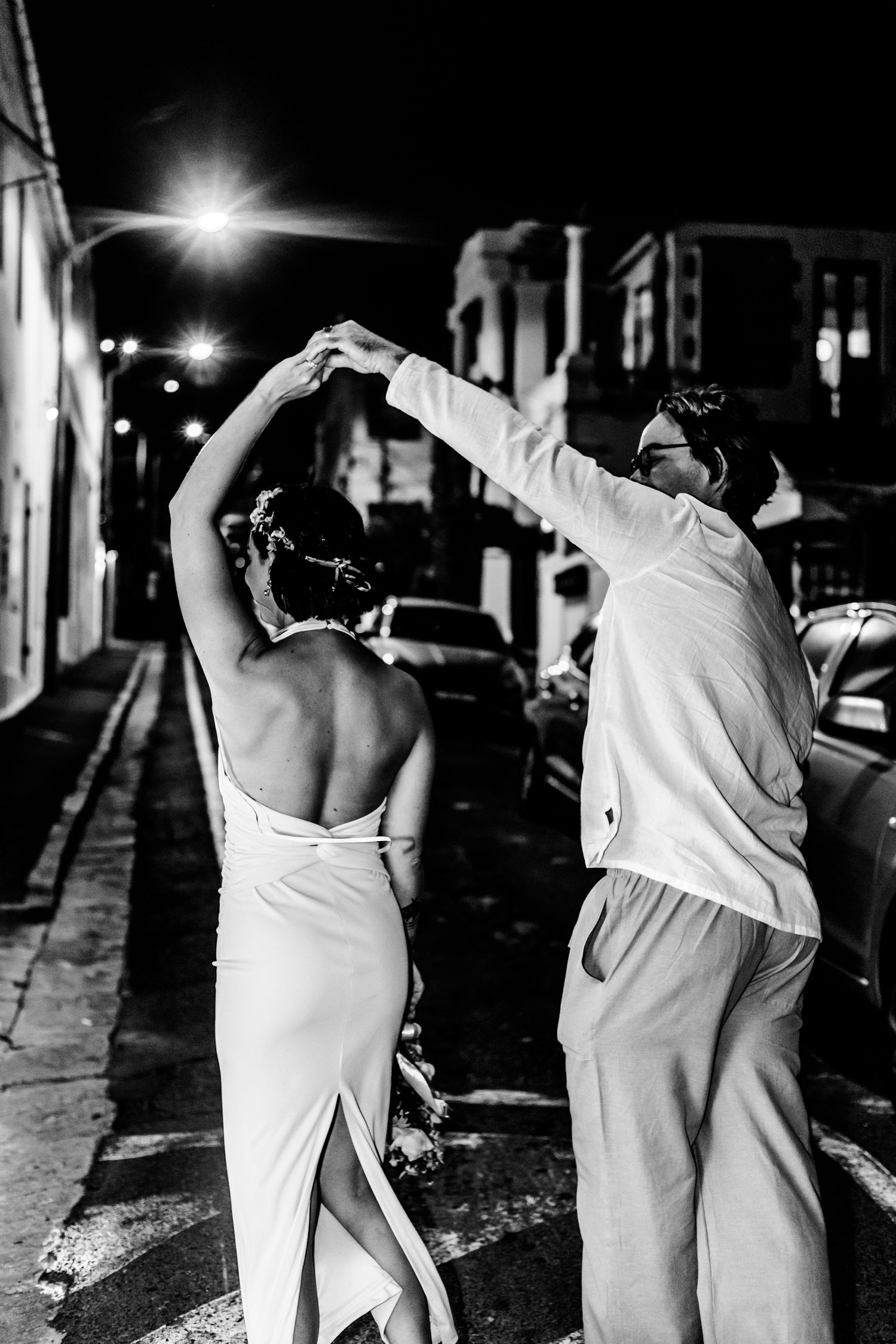 A couple in white attire dancing together on a nighttime street under street lights.