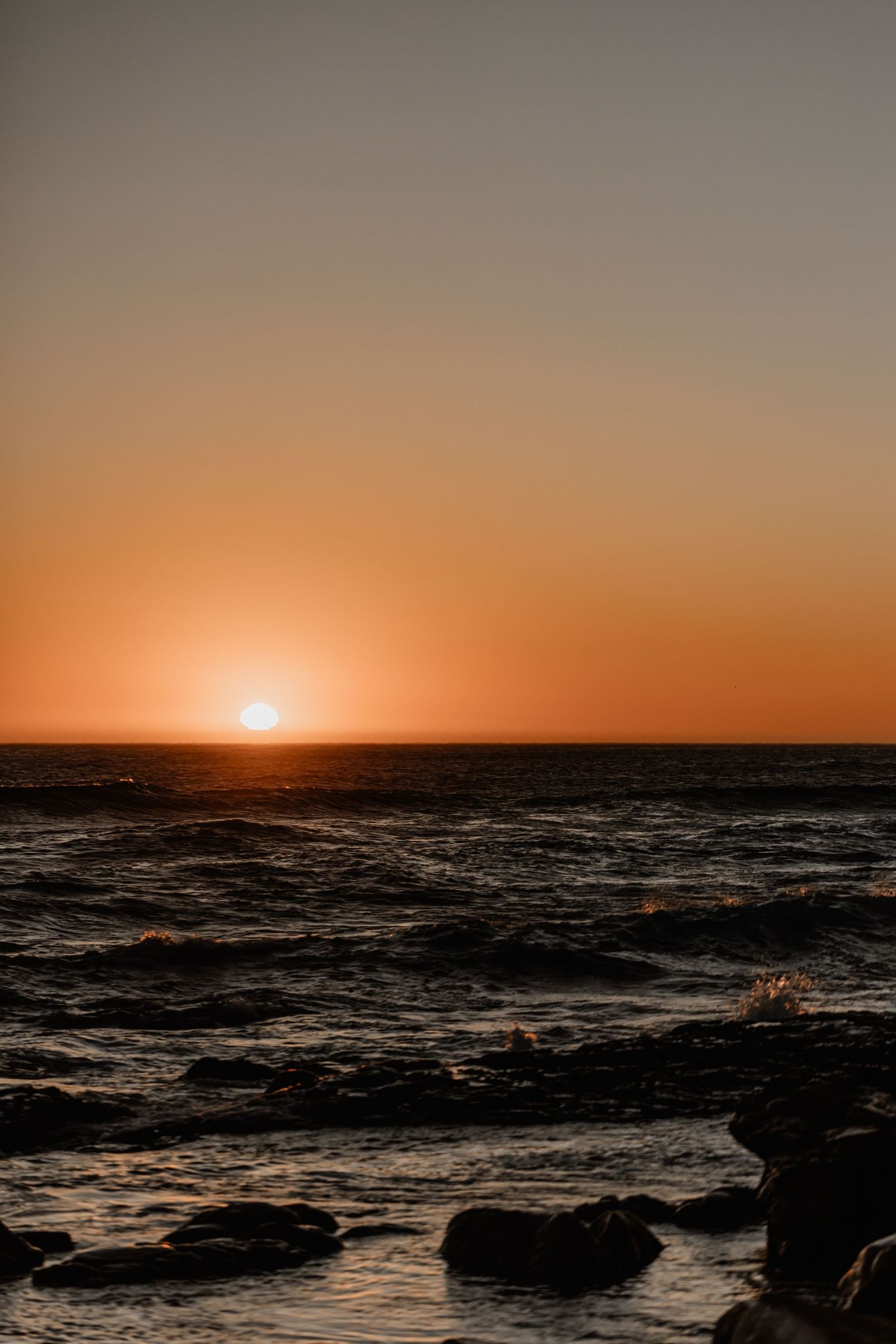A dramatic sunset over the ocean with rocky coastline silhouetted against an orange sky and rippling waves.